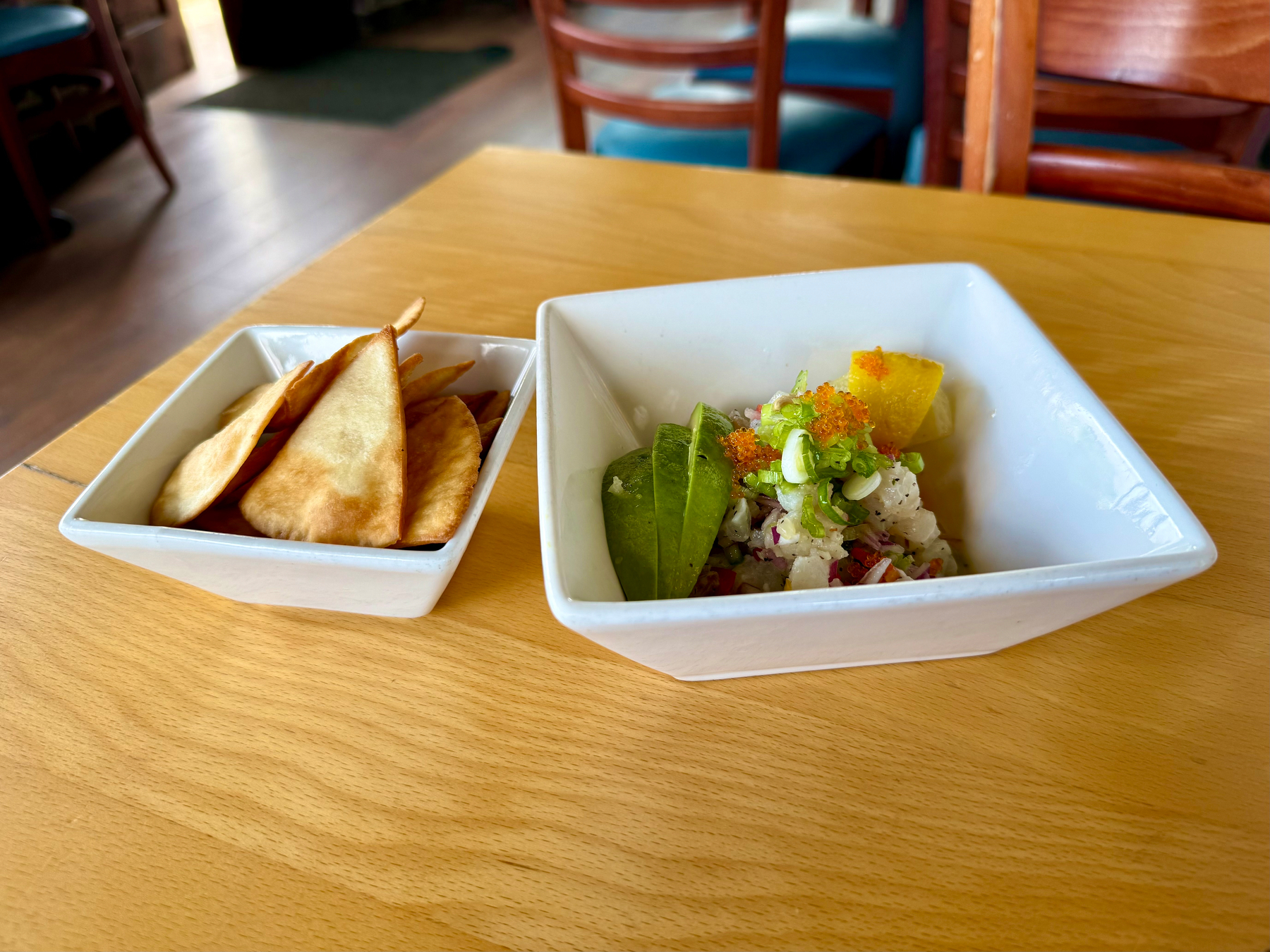 A square white bowl filled with ceviche sits on a light wooden table in a restaurant. The ceviche contains diced white fish, finely chopped red onion, green onions, and orange fish roe, garnished with two slices of avocado and a lemon wedge. Next to it is another square white bowl containing several golden-brown triangular fried tortilla chips. In the background, there are wooden chairs with teal seats and a dimly lit interior with a doorway leading outside.