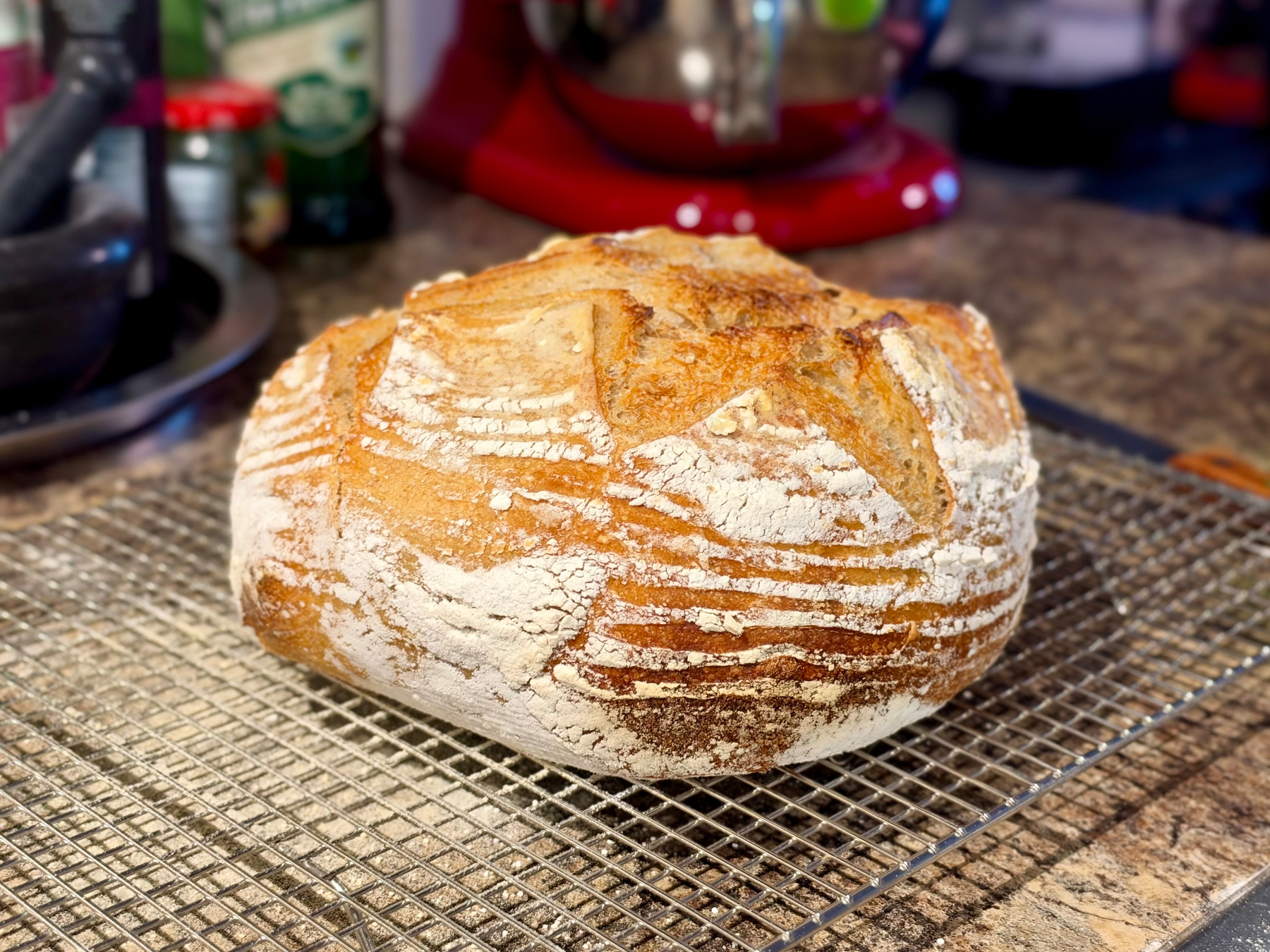 A freshly baked round loaf of artisan bread rests on a metal cooling rack in a kitchen. The bread has a golden-brown crust with a rustic pattern of flour-dusted lines and ridges, likely from being proofed in a banneton basket. The top is slightly cracked with a beautiful star-like scoring pattern, revealing a well-baked crust. The background shows a countertop with kitchen items including a mortar and pestle, jars, a bottle of oil, and a red stand mixer, all slightly out of focus.