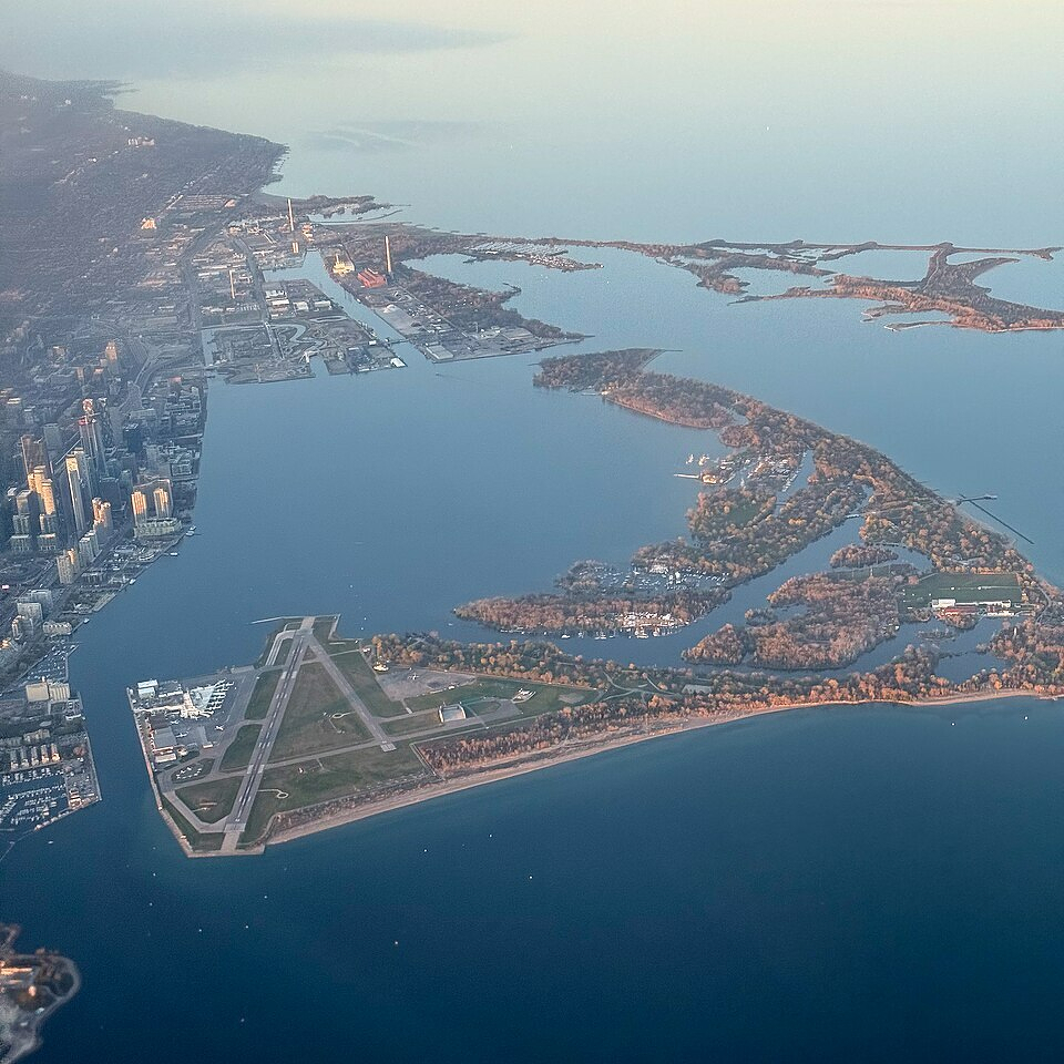 An aerial view of Toronto’s waterfront and the Toronto Islands during daylight. In the foreground, Billy Bishop Toronto City Airport is clearly visible, with its triangular runway layout surrounded by water. The Toronto Islands, covered with a mix of trees and open clearings, extend into Lake Ontario, with lagoons and inlets carving through them. Several small boats and docks are visible along the island’s shorelines. The downtown Toronto skyline appears on the left side, with clusters of tall buildings, while the industrial port lands stretch into the distance along the water. The calm lake reflects the soft light of early morning or late afternoon, and the horizon fades into a hazy sky.
