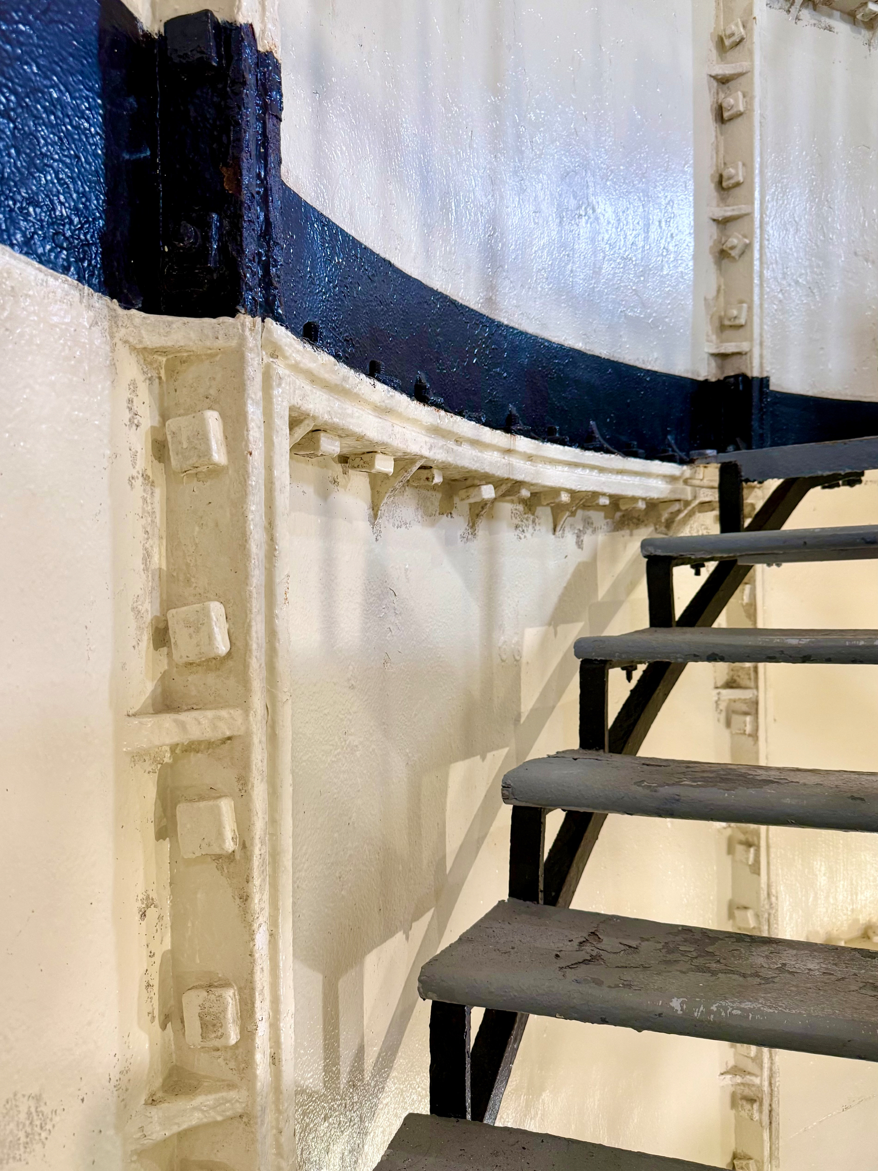 A close-up view of a metal spiral staircase inside a tall structure with cream-coloured walls and a horizontal dark blue stripe running across the curved surface. The staircase steps are painted grey and show signs of wear and chipping. The supporting metal frame is black, and the walls feature protruding rivets and square bolts arranged in vertical and horizontal lines. The bright lighting highlights the glossy texture of the painted surfaces and casts defined shadows of the stair steps on the wall.