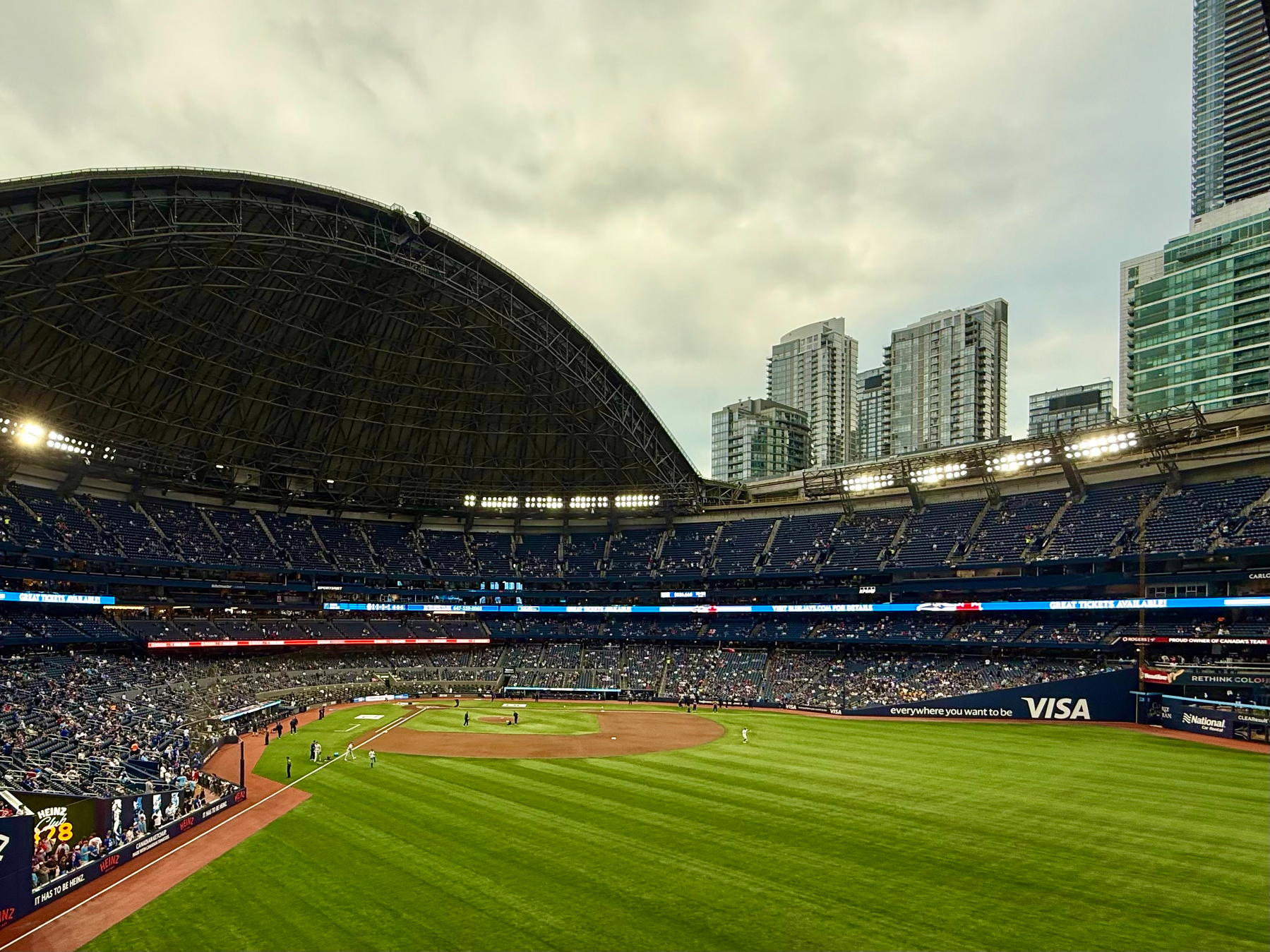 A wide view of a baseball stadium with a partially open retractable roof under a cloudy sky. The field is green with players positioned near the infield, and the stands are sparsely populated with spectators. Tall modern buildings rise in the background beyond the stadium. Bright stadium lights illuminate the seating areas, and electronic advertising boards line the outfield wall, featuring a large blue VISA sign and other ads along the field’s edge.