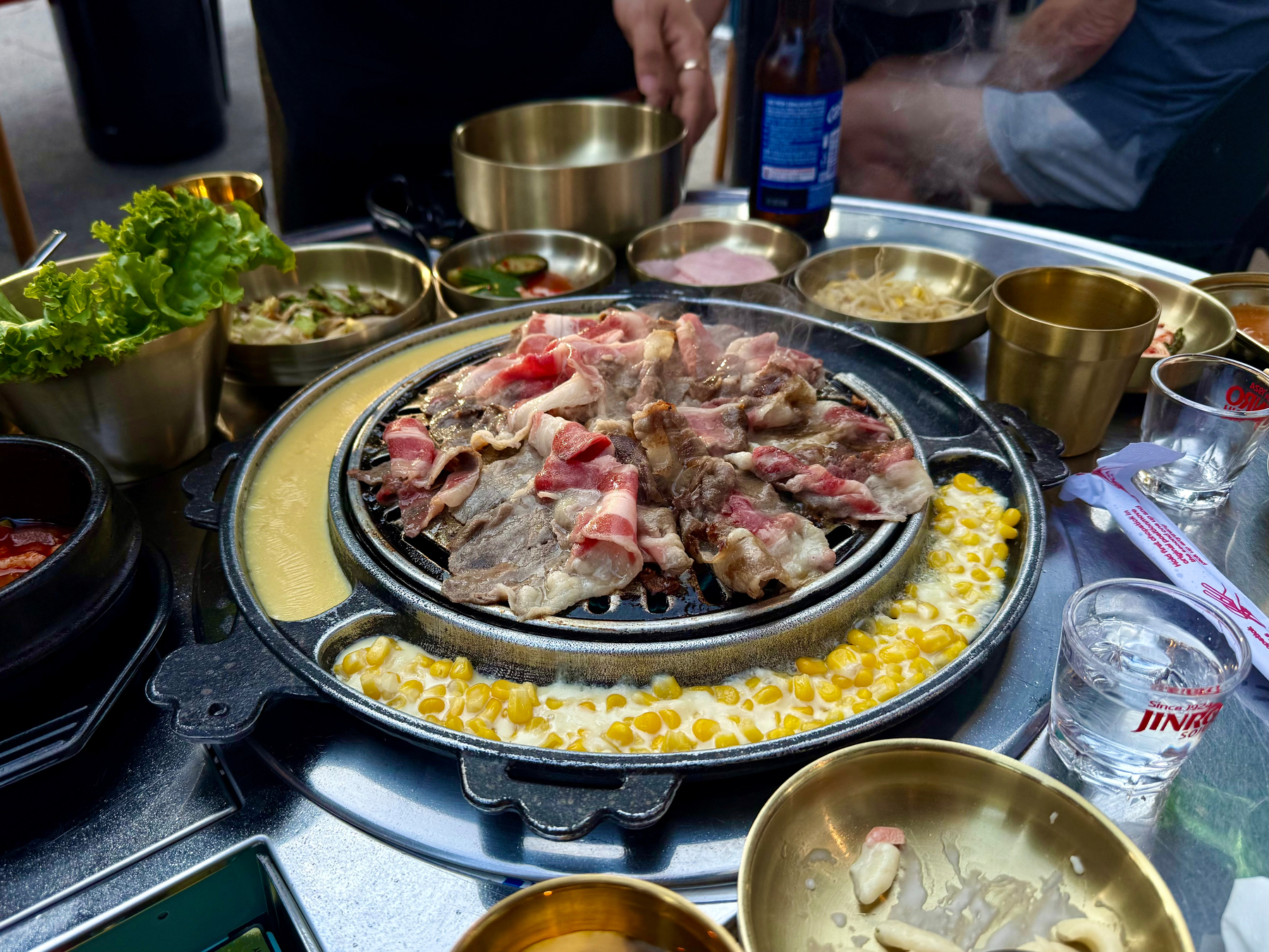 A Korean barbecue setup on a metal table with various dishes and side plates. In the centre, slices of raw and partially cooked beef are grilling on a round tabletop grill surrounded by a ring containing melted cheese and corn. Steam rises from the grill, and several golden metal bowls and cups are scattered around, filled with side dishes like lettuce, bean sprouts, and other vegetables. A black stone pot with a red stew sits on the side, and a glass of clear soju along with another empty glass are on the table. A blue-labelled beer bottle and a wrapped disposable chopstick set are also visible. People’s hands and part of an arm can be seen in the background.