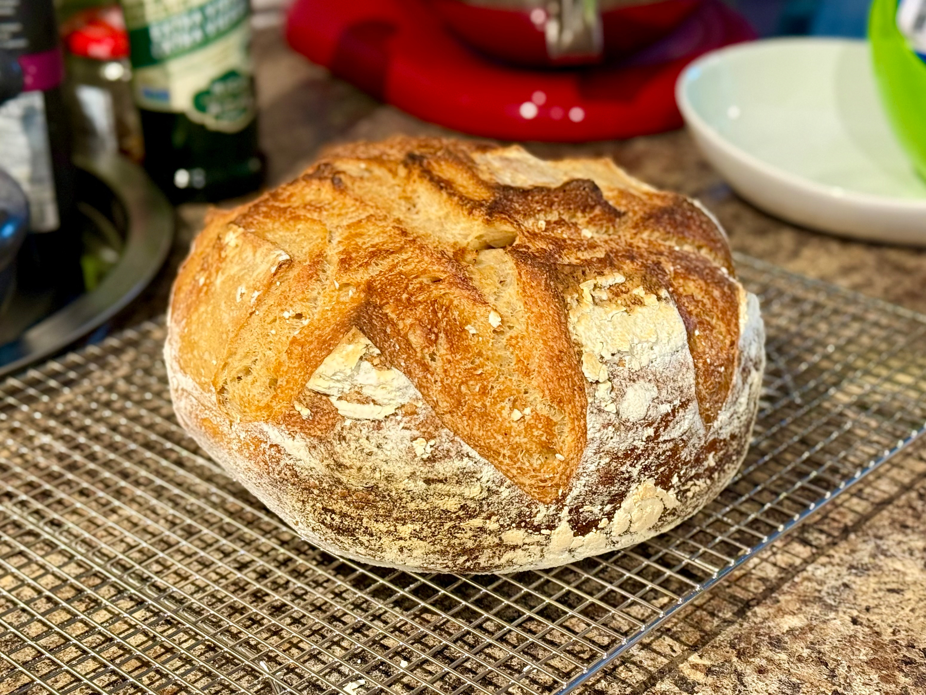 A freshly baked round loaf of artisan bread with a golden-brown crust sits on a wire cooling rack on a kitchen counter. The crust is rustic with deep, uneven scoring patterns and patches of white flour. The bottom edge of the loaf appears slightly darker and crisp. In the background, there is a red stand mixer, a white bowl, a green bowl, and various kitchen items including a dark sink and bottles, one of which is green. The counter surface is speckled brown, and the lighting highlights the bread’s texture and colour.