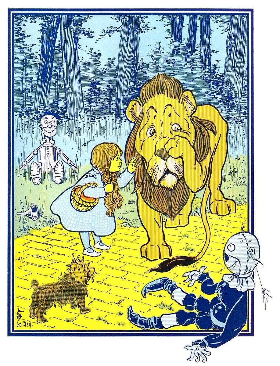 Illustration in a blue-bordered frame of a woodland scene from *The Wonderful Wizard of Oz*. Tall trees with blue-shaded bark rise against a pale blue sky. A yellow brick road runs through the foreground. Dorothy, a young girl with long braids, wears a blue gingham dress and white shoes and carries a yellow woven basket on her arm. She looks up at the Cowardly Lion and points at him. The lion is large and golden-yellow, with a shaggy mane and a tearful, worried expression, and he raises a paw to his face. Toto, a small brown shaggy dog, stands on the road in the lower left. In the background on the left, the silver Tin Woodman stands smiling among the grass, with a small oil can lying near his feet. In the lower right, the Scarecrow lies sprawled across the brick road in blue clothing, his stitched face turned upward and straw showing at his hands and neck. A small signature, reading "Den," appears in the lower left corner.
