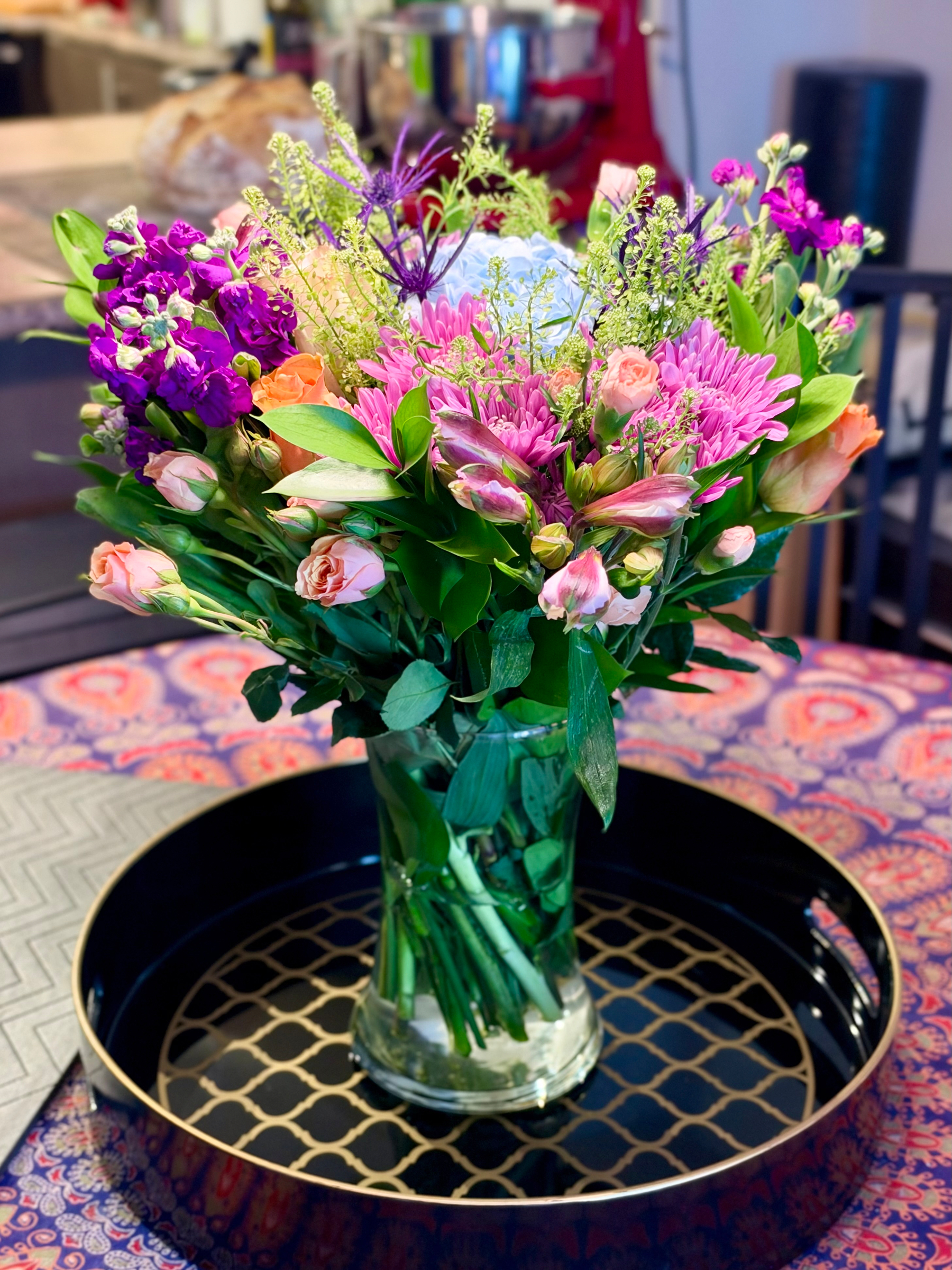 A vibrant bouquet of assorted flowers in a clear glass vase sits on a decorative round tray with a black base and gold lattice pattern. The bouquet features a mixture of pink roses, peach roses, purple stock flowers, pink and white alstroemeria, bright pink chrysanthemums, green filler foliage, and small clusters of purple thistle-like blooms. The flowers are lush and arranged in a dome shape, with green leaves and stems visible through the water in the vase. The tray rests on a table covered with a colourful paisley-patterned tablecloth in shades of purple, pink, and orange. In the background, a kitchen counter with a loaf of bread and a silver stand mixer is visible, along with a black chair and part of a dark cylindrical object.