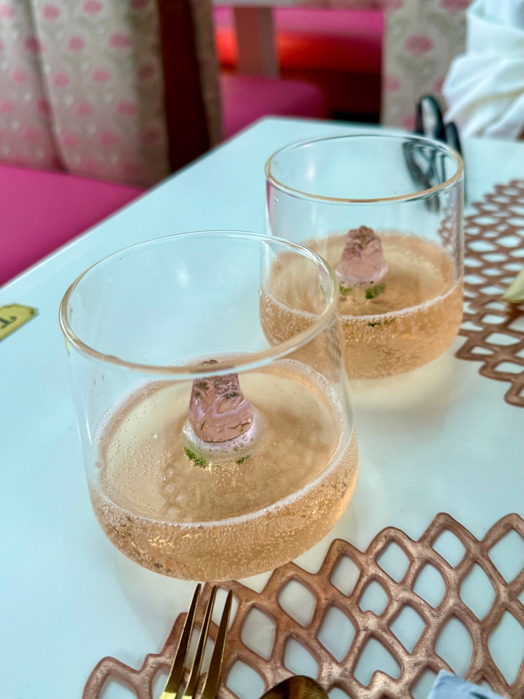 Two clear glasses filled with a light pink bubbly drink sit on a white table. Each glass contains a pink, bear-shaped ice cube with a hint of green mint at the base. The table is decorated with a rose-gold lattice-patterned placemat, and a gold fork and spoon are partially visible in the foreground. In the background, there are pink cushioned seats with a subtle floral pattern, and part of a white cloth and black-handled scissors are visible to the right.