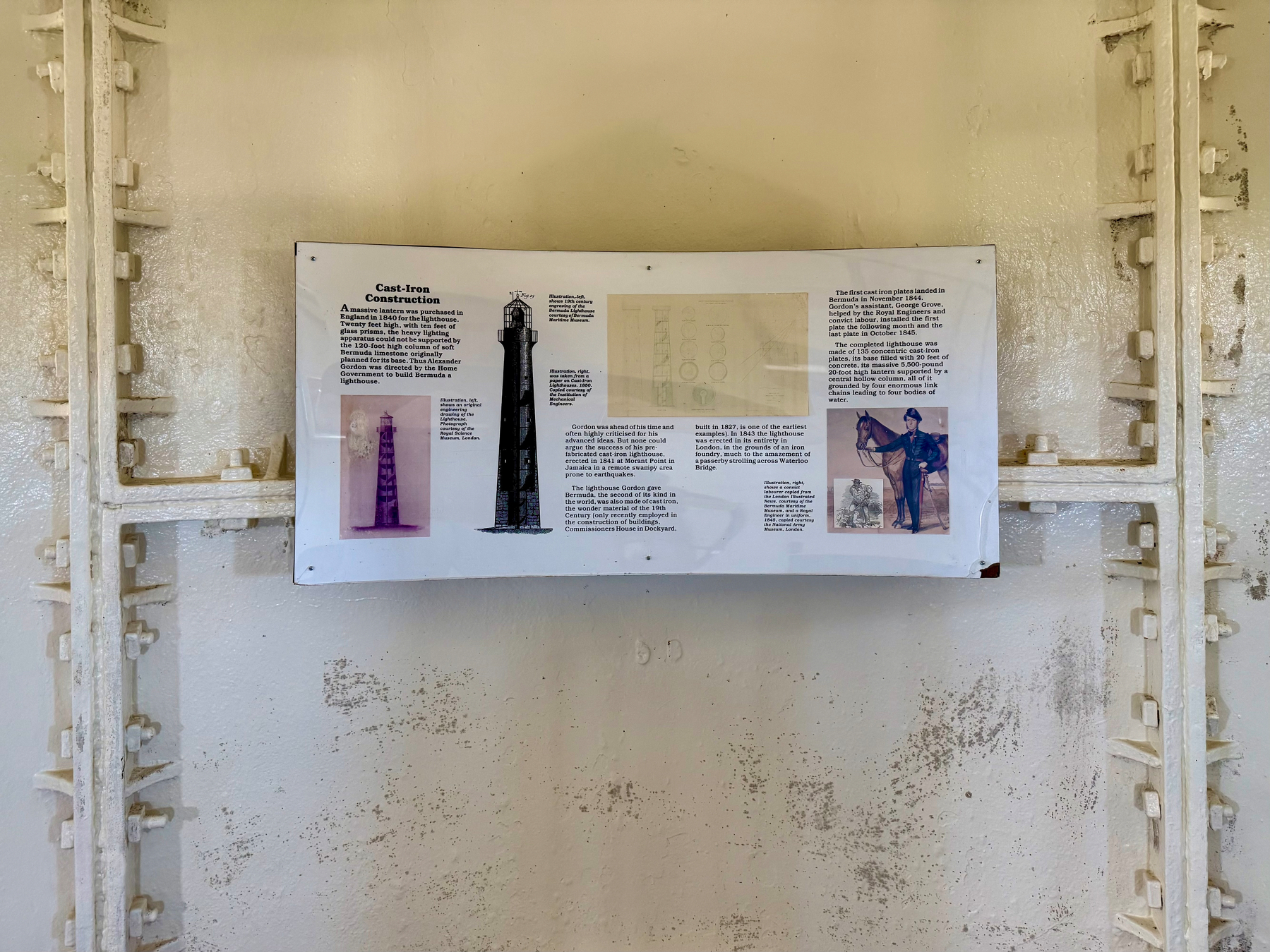 A rectangular informational sign is mounted on a cream-coloured wall with visible metal structural supports painted white, featuring rivets and crossbars. The sign is titled “Cast-Iron Construction” and describes the construction of a lighthouse using cast iron. On the left side, there is a purple-toned illustration of a lighthouse and a black-and-white drawing of the lighthouse structure. In the centre, there is a faint diagram or blueprint of the lighthouse’s design. On the right, there is text describing the history and completion of the lighthouse, accompanied by an image of a man in historical military attire holding a horse and a small illustration beneath it. The surrounding wall shows areas of weathering, with patches of peeling or stained paint.
