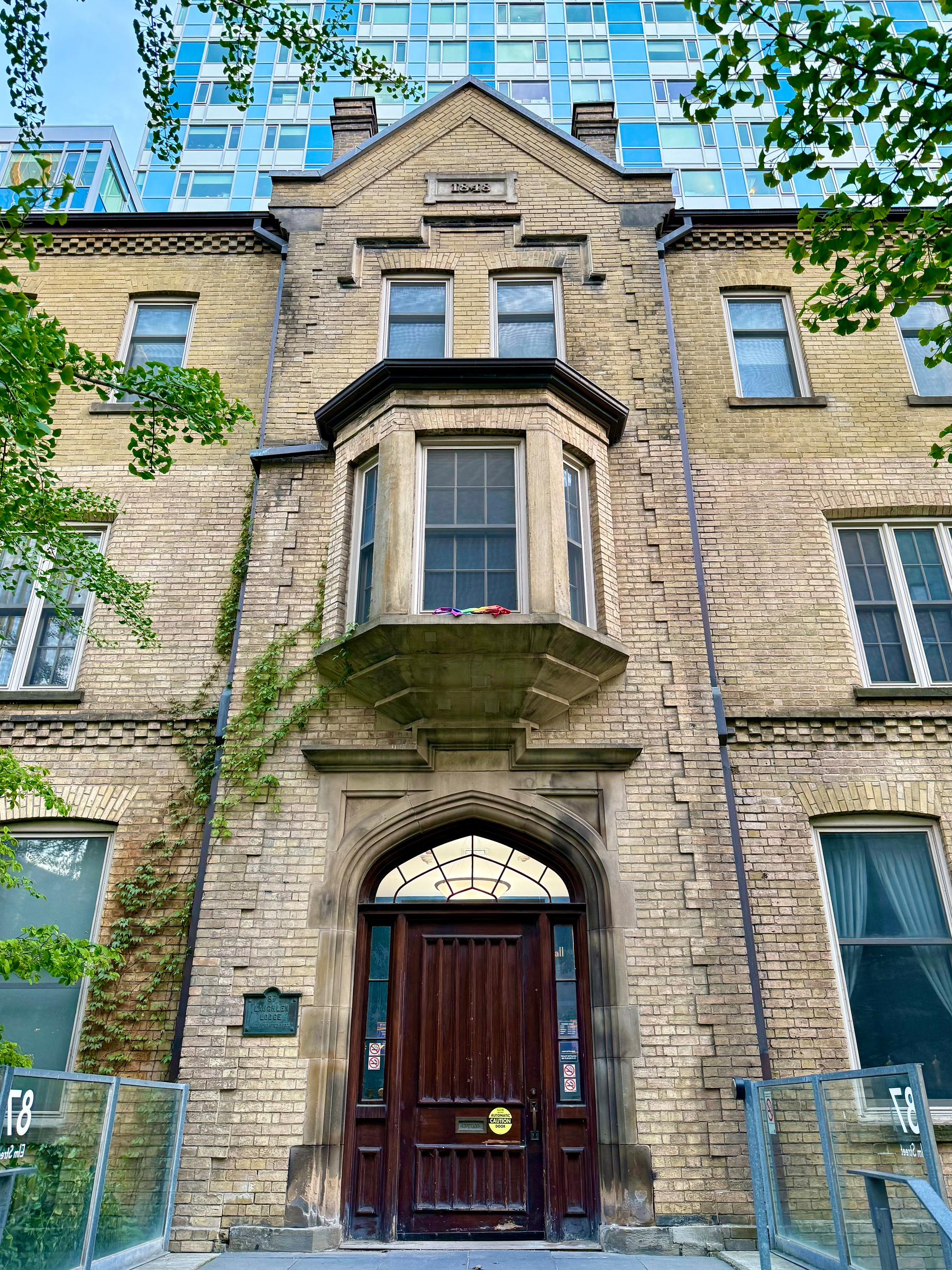 A historic brick building with a central arched wooden door and a large window above it, flanked by two tall rectangular windows. The upper façade features a bay window with stone detailing and three smaller windows above, beneath a triangular gable with the year “1893” engraved. Vines partially climb the left side of the building. The structure is fronted by modern glass railings, and a tall glass high-rise towers in the background. Green tree branches frame the scene from the top left and right corners.