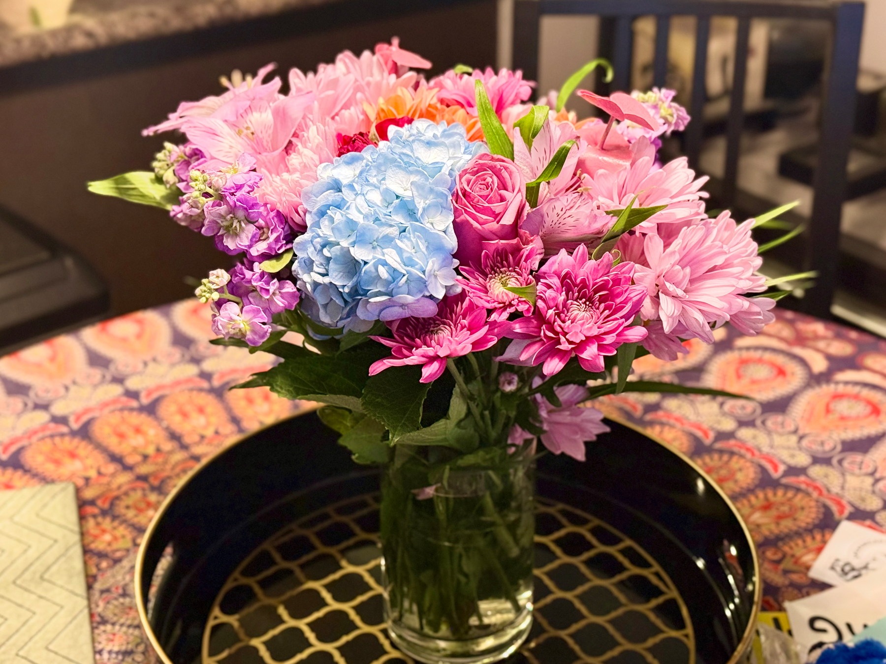 A clear glass vase holds a vibrant bouquet of flowers, displayed on a black round tray with a golden lattice pattern. The bouquet includes a variety of colourful blooms: light blue hydrangeas, pink roses, pink and purple chrysanthemums, light pink lilies, and small clusters of purple flowers. The flowers are lush with green leaves interspersed throughout. The background shows a patterned tablecloth in shades of red, blue, and orange with a paisley design, and part of a black chair and a countertop. On the left side of the table, there is a folded zigzag-patterned napkin in light colours, and on the right, some papers and a small blue flower are partially visible. The scene is warmly lit, highlighting the vivid colours of the flowers.