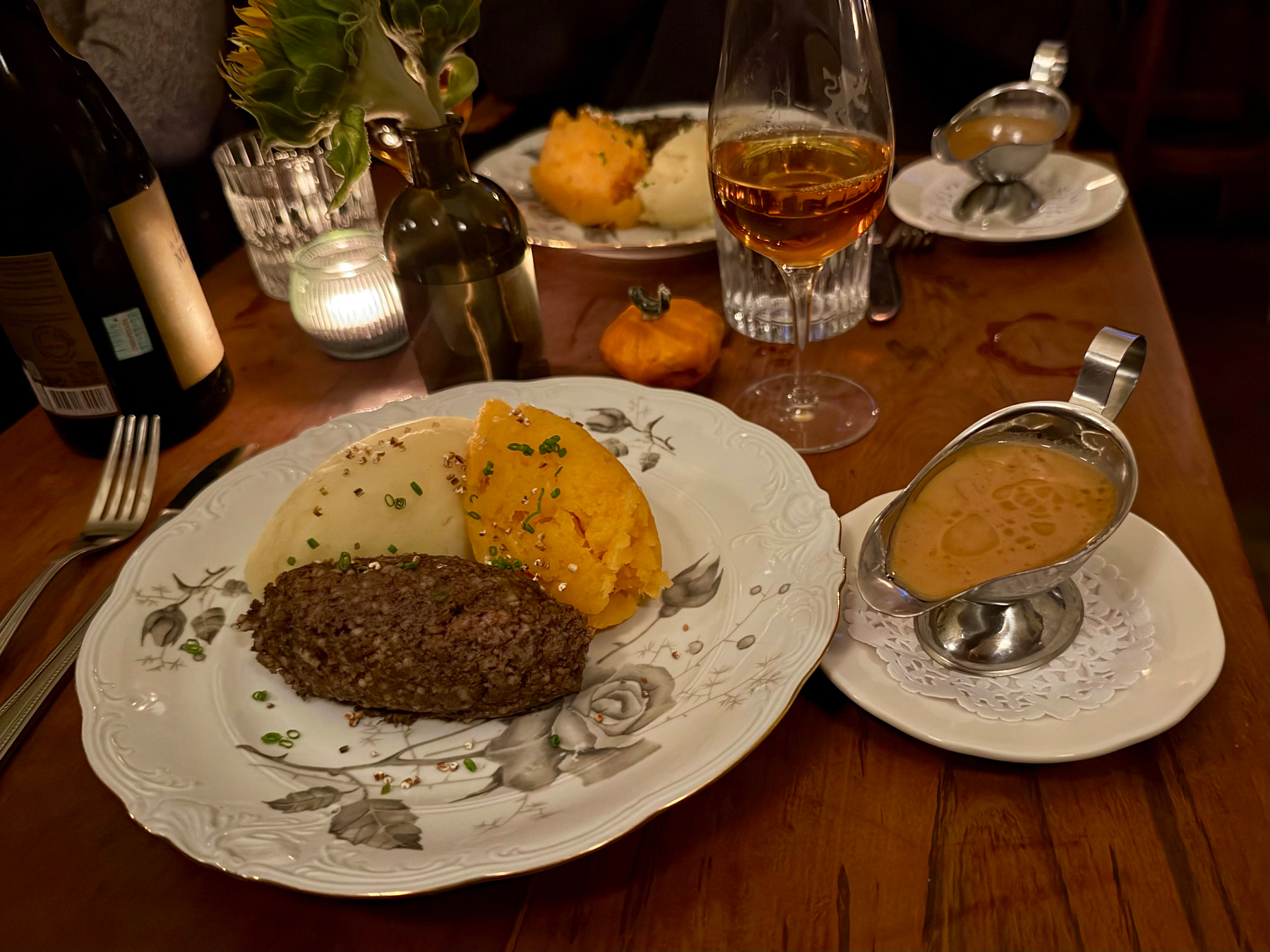 A warmly lit dining table with a plate of food featuring a serving of mashed potatoes, mashed turnip or carrot, and a portion of haggis garnished with chopped chives. On the right side of the plate is a small metal gravy boat filled with light brown gravy, resting on a white doily on a small plate. A wine glass filled with amber-coloured liquid and a glass of water are in the background, along with a second plate of food featuring similar mashed vegetables. A small orange decorative gourd and a lit candle in a ribbed glass holder add to the cozy ambiance. A dark wine bottle, a fork, and a glass vase holding a greenish-yellow flower are also on the wooden table. The overall setting evokes a warm, intimate dining experience.