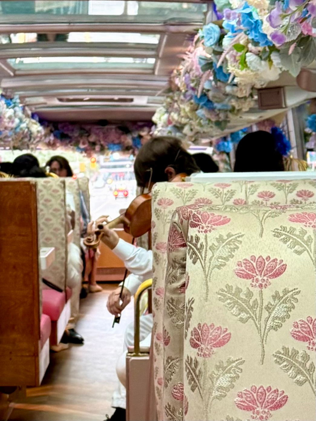 The image shows the interior of a decorative bus or tram, with floral-patterned seat covers in cream, pink, and green. The ceiling and upper walls are adorned with abundant artificial flowers in shades of blue, pink, purple, and white. The bus has large windows and a glass roof, letting natural light in. In the middle of the aisle, a person dressed in white is playing a violin, with other passengers seated and some visible in the background. The floor appears wooden, and the overall atmosphere is bright and elegant. Traffic lights and blurred city elements can be seen through the windows at the front.