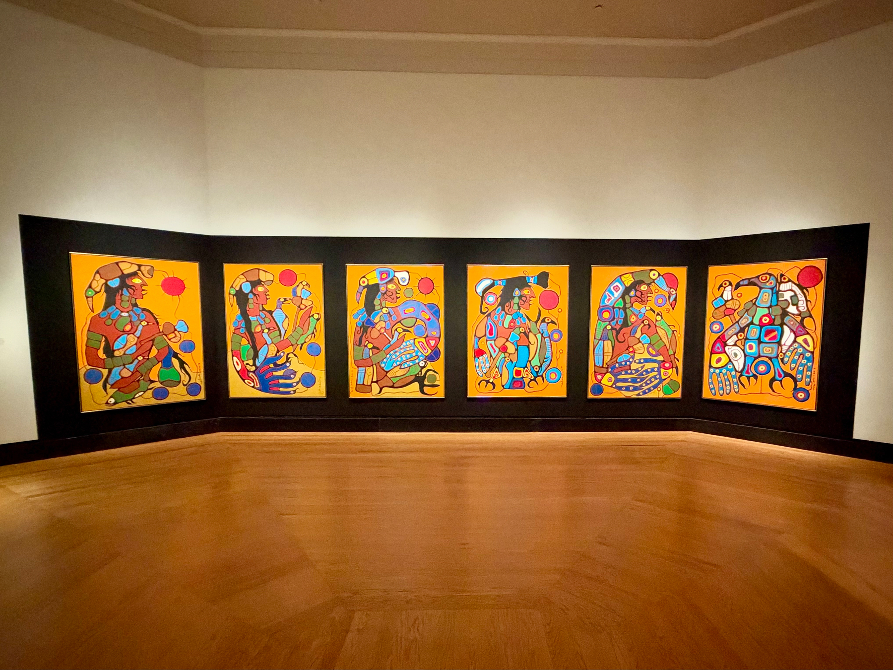 Seven colorful, abstract paintings with human and animal figures are displayed on a connected black panel in a gallery with wooden flooring.