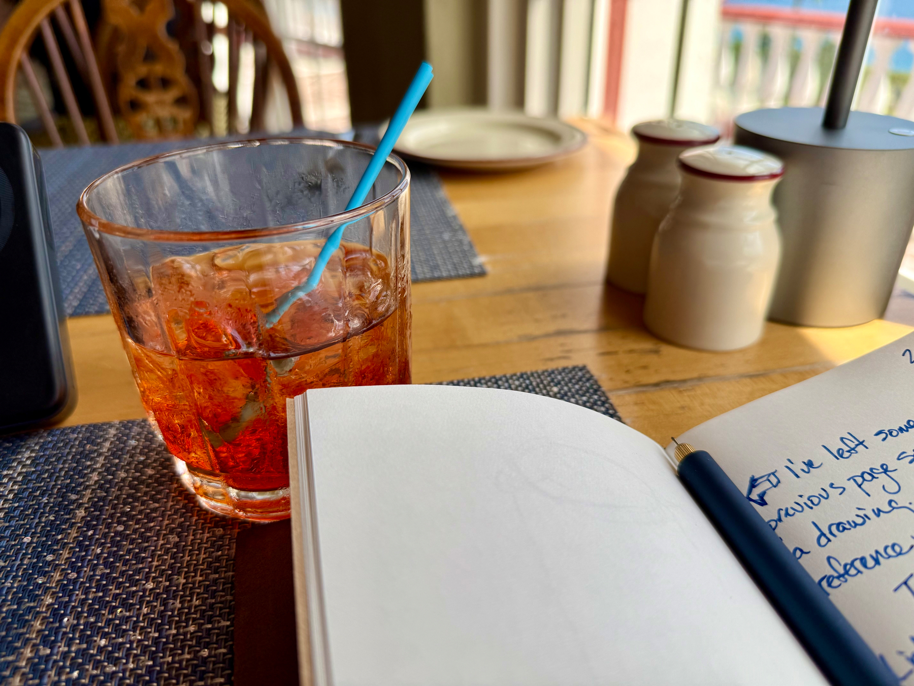 A wooden table set for a meal with a blue woven placemat, a glass filled with an amber-coloured drink and ice cubes, and a light blue straw. In the foreground, an open notebook with a dark blue pen rests on the table, showing handwritten notes in blue ink. Behind the glass is a beige plate, and two cream-coloured ceramic shakers with red rims sit near a modern silver lamp. Wooden chairs and a balcony railing with a view of the outdoors are blurred in the background, letting in natural daylight.