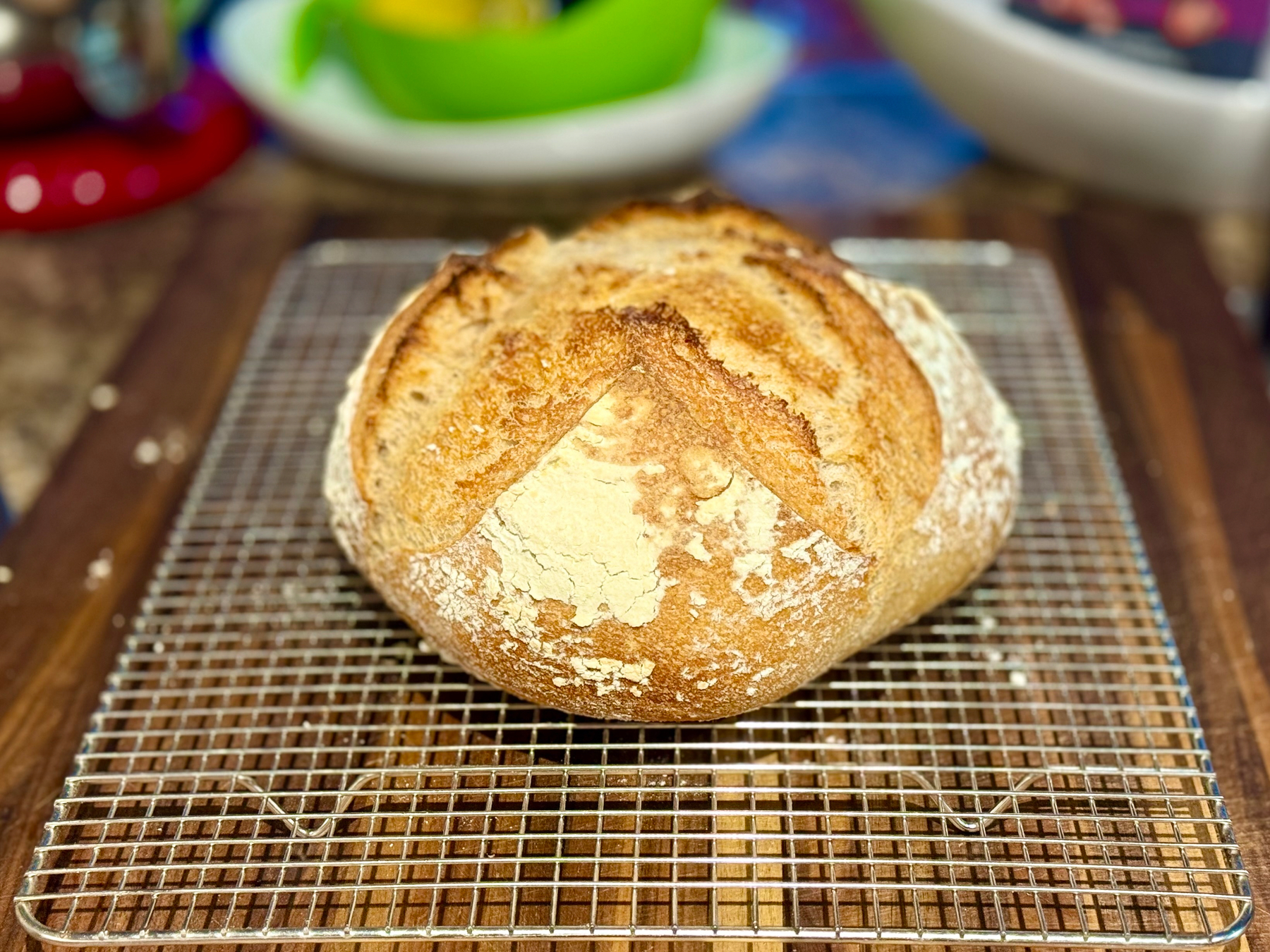 A freshly baked round loaf of artisan bread with a golden-brown crust rests on a metal cooling rack atop a wooden surface. The bread has a rustic appearance with a cross-shaped score on top and a dusting of flour, particularly on the sides. The loaf is well-risen, with deep ridges where the scoring expanded during baking. In the blurry background, colourful kitchen items are visible, including a green bowl, a white plate, and a red appliance.
