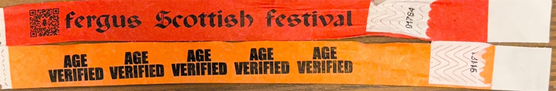 Two coloured wristbands are laid out flat on a wooden surface. The top wristband is bright red with a QR code on the left and the words “fergus Scottish festival” printed in a bold, black medieval-style font. The end of the wristband has a white adhesive strip with the handwritten number “01784.” The bottom wristband is orange with the words “AGE VERIFIED” repeated multiple times in bold black lettering. Its white adhesive strip also has a handwritten number, “31746.”