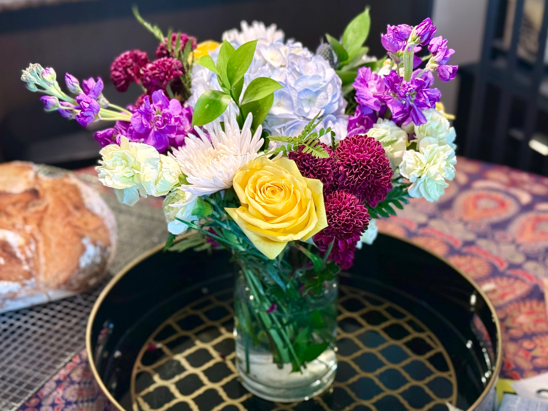 A glass jar filled with a colourful flower arrangement sits on a black and gold patterned tray. The bouquet includes a prominent yellow rose, light purple hydrangea, bright purple stock flowers, deep burgundy pompom chrysanthemums, white carnations, and green foliage. Behind the tray on the left is a loaf of rustic bread dusted with flour, resting on a textured mat. The background includes a patterned tablecloth with orange, red, and purple designs, and dark furniture is slightly out of focus.
