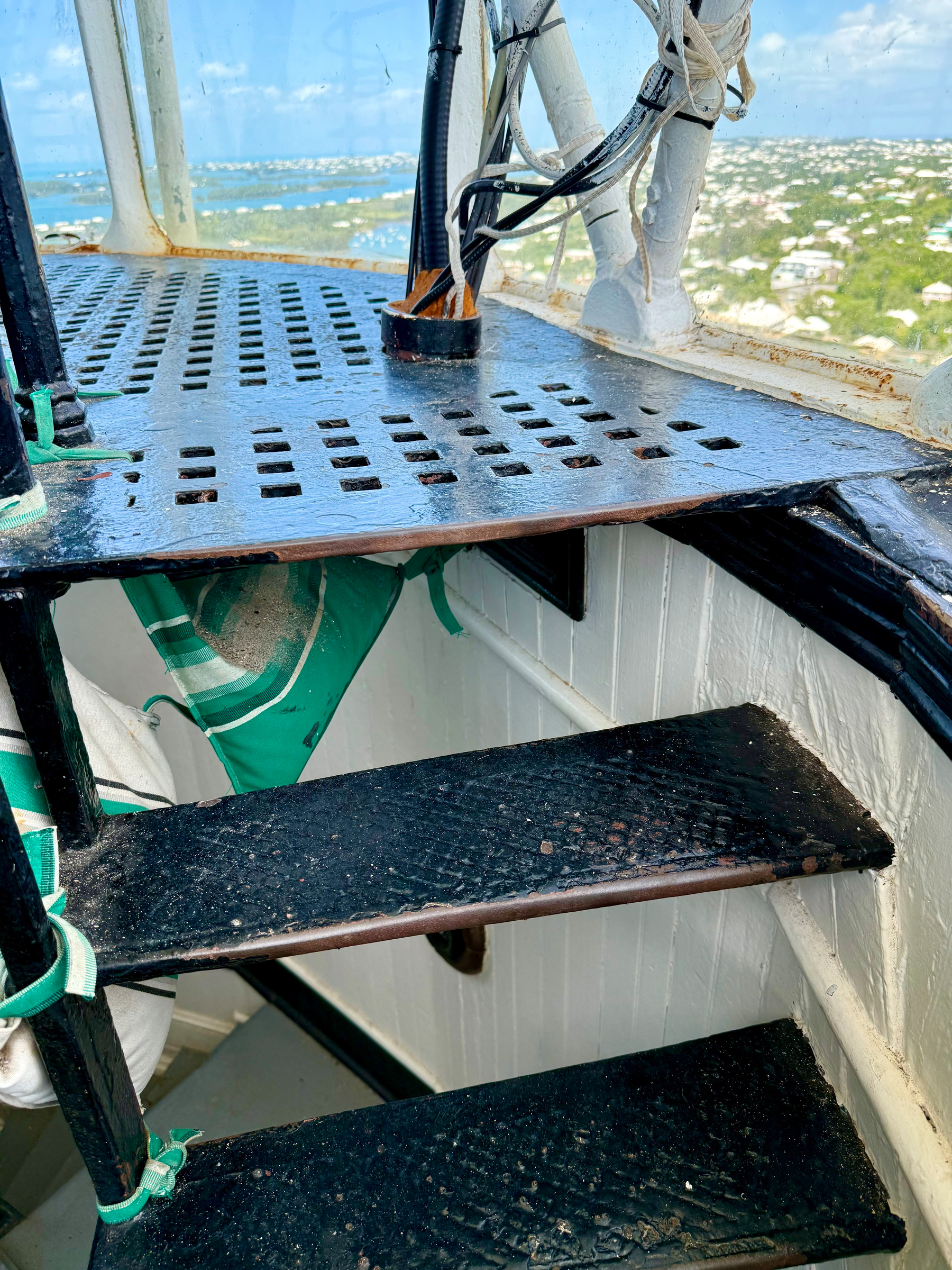 A close-up view of a black metal spiral staircase inside a structure, possibly a lighthouse. The steps are worn and textured with signs of rust and chipping paint. Green straps secure white cylindrical objects, likely life preservers, to the railing. Above the steps, a perforated metal platform with square holes supports several bundled cables and wires wrapped in white tape, leading upward through a black circular fitting. The surrounding walls are white with black trim, and the windows reveal a panoramic view of a coastal town with blue water, green land, and scattered buildings under a bright sky with soft clouds.