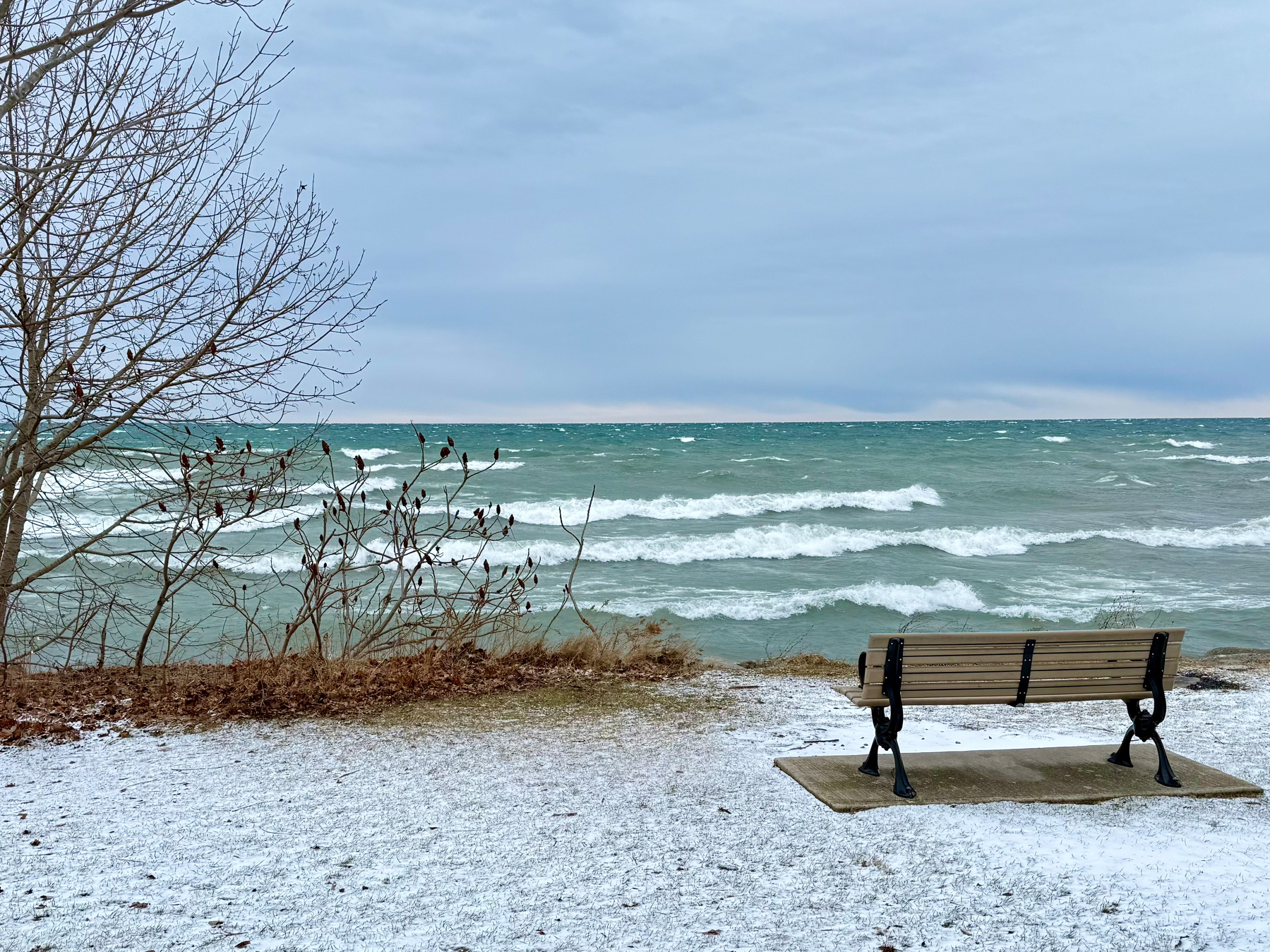 A wooden bench is situated on a snowy patch of ground overlooking a wavy, turquoise lake. On the left, leafless tree branches and shrubs frame the scene under a cloudy sky. The setting conveys a serene and chilly lakeside atmosphere.