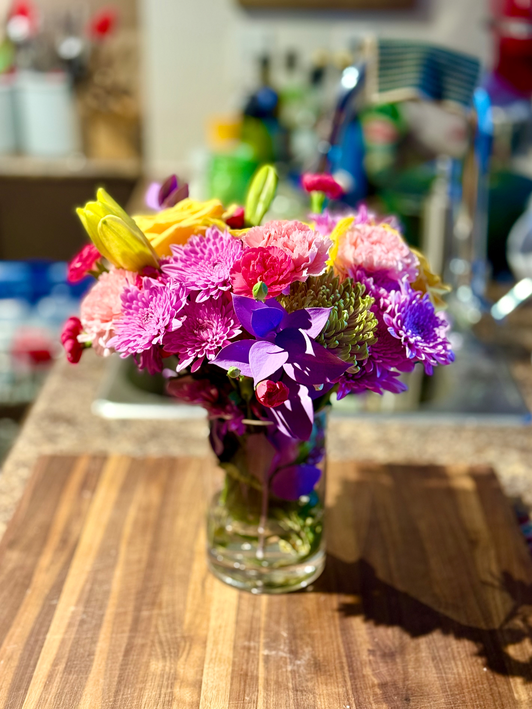 A vibrant bouquet of flowers in a clear glass vase sits on a wooden countertop. The arrangement features a variety of blooms, including bright yellow lilies, soft pink carnations, purple chrysanthemums, small red buds, and clusters of green button mums. Some deep purple leaves and petals are interspersed throughout the bouquet, adding contrast. The stems are visible through the water in the vase. Behind the flowers, a kitchen sink and counter with various items—including a faucet, a striped sponge, bottles, and blurred kitchen tools—can be seen in the background.