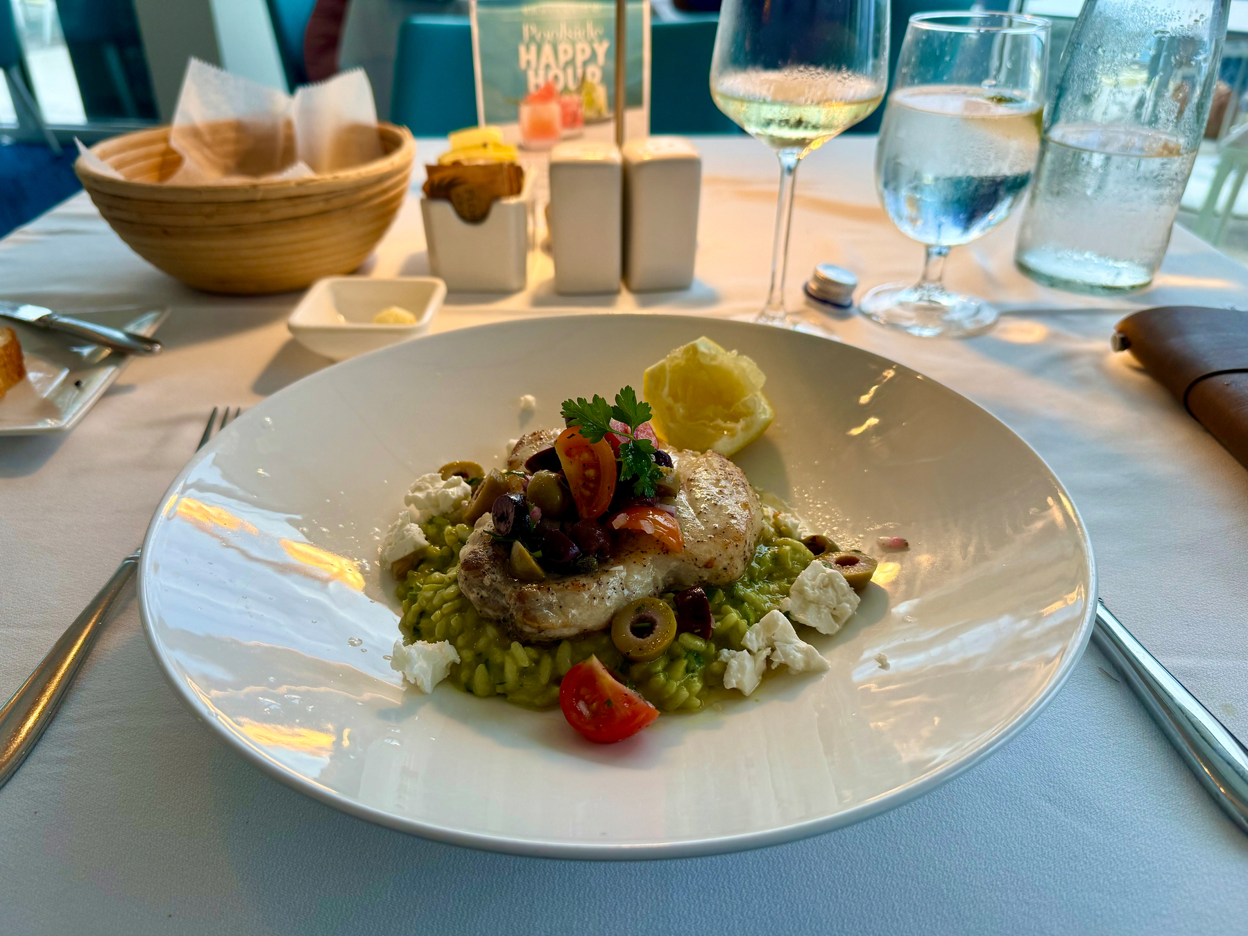 A plated gourmet dish is served on a white tablecloth in a fine dining setting. The dish features a seared piece of fish topped with a garnish of halved cherry tomatoes, sliced green and black olives, and a sprig of parsley. It rests on a bed of creamy green risotto, surrounded by chunks of white cheese and a small yellow garnish resembling a flower. In the background, there is a wicker bread basket with parchment paper, a small square dish of butter, a rectangular holder for condiments, a glass of white wine, a glass of water, and a bottle of water with condensation. Reflections of light are visible on the glossy plate.