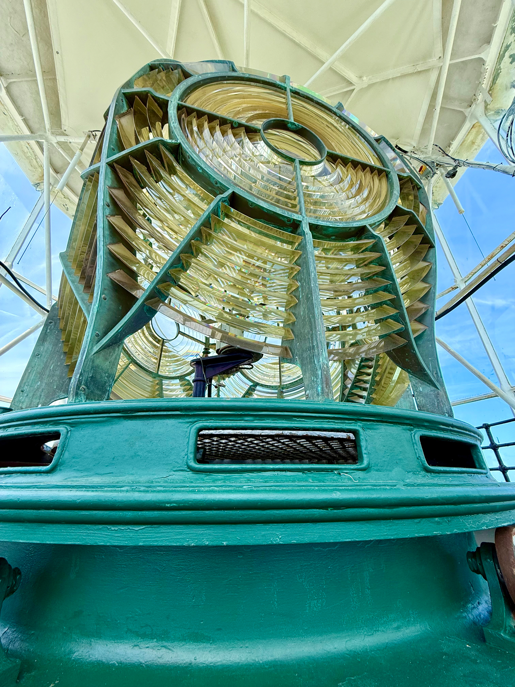 A close-up view of the large Fresnel lens inside a lighthouse, seen from a low angle. The lens features multiple concentric glass panels with a golden hue, framed by a green metal structure that shows signs of weathering. The lens housing sits atop a sturdy green-painted metal base with rectangular vents and a grated opening. Behind the lens, the circular glass enclosure of the lighthouse is visible, with white metal support beams and a bright blue sky in the background.