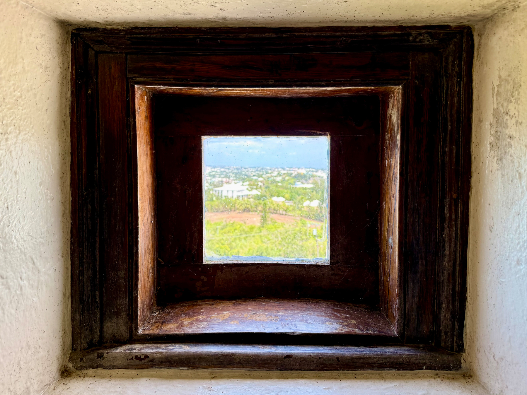 A small square window with a deep, dark wooden frame is set into a thick, white plastered wall. The frame has a tiered, beveled design that creates a tunnel-like effect, drawing the eye to the view outside. Through the glass, a sunlit landscape with green fields, scattered white buildings, and a hint of blue sky and ocean in the distance is visible. The contrast between the dark wood, white walls, and bright outside scenery is striking.