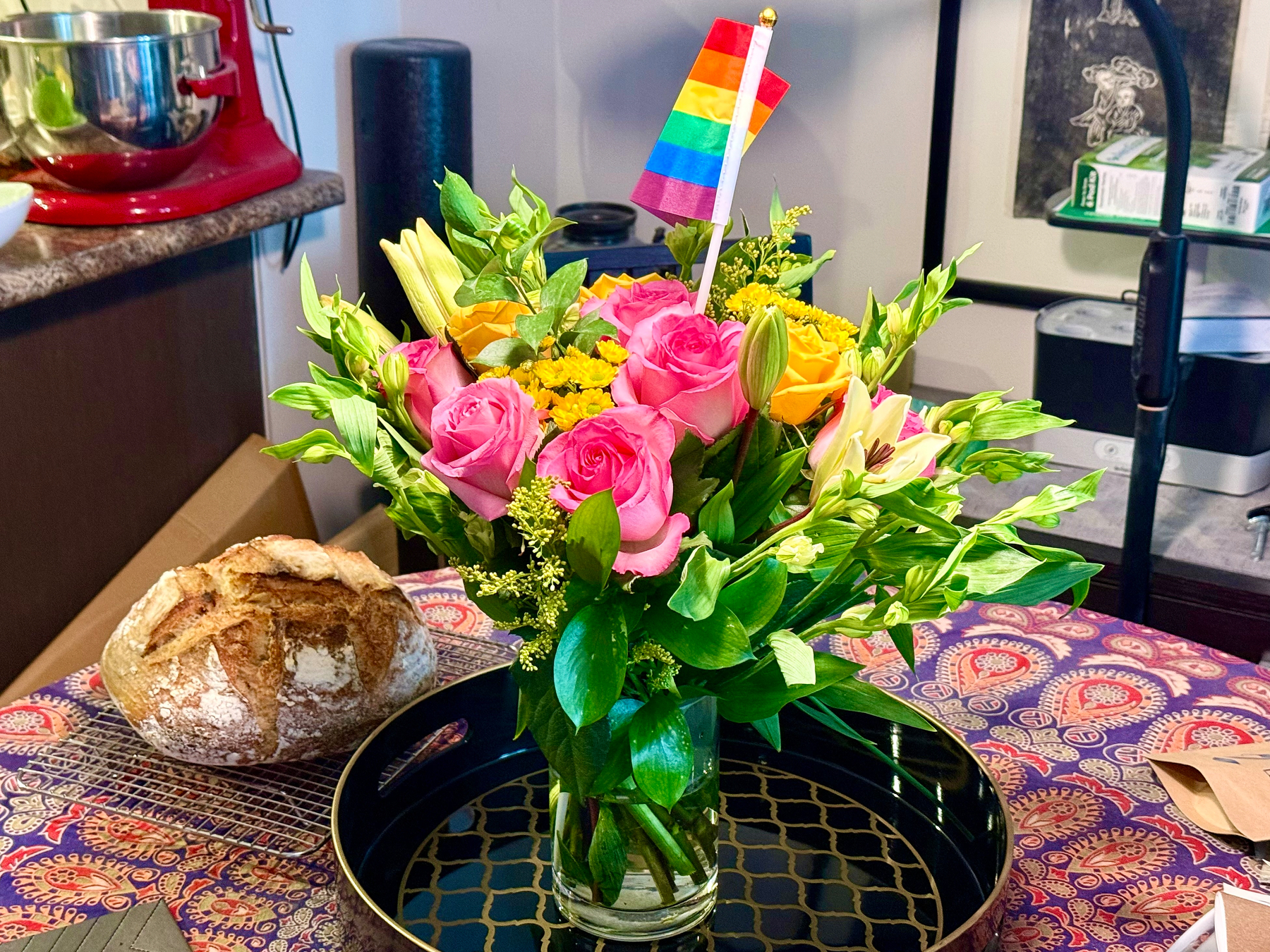 A colourful bouquet of flowers in a glass vase sits on a black tray with a gold geometric pattern, placed on a table covered with a vibrant, patterned tablecloth. The flowers include pink roses, yellow lilies, small yellow flowers, and various green leaves. A small rainbow Pride flag is placed in the bouquet. To the left, there is a round loaf of rustic bread cooling on a wire rack. In the background, a red stand mixer, a black foam roller, and part of a kitchen counter are visible, along with some boxes and kitchen items.