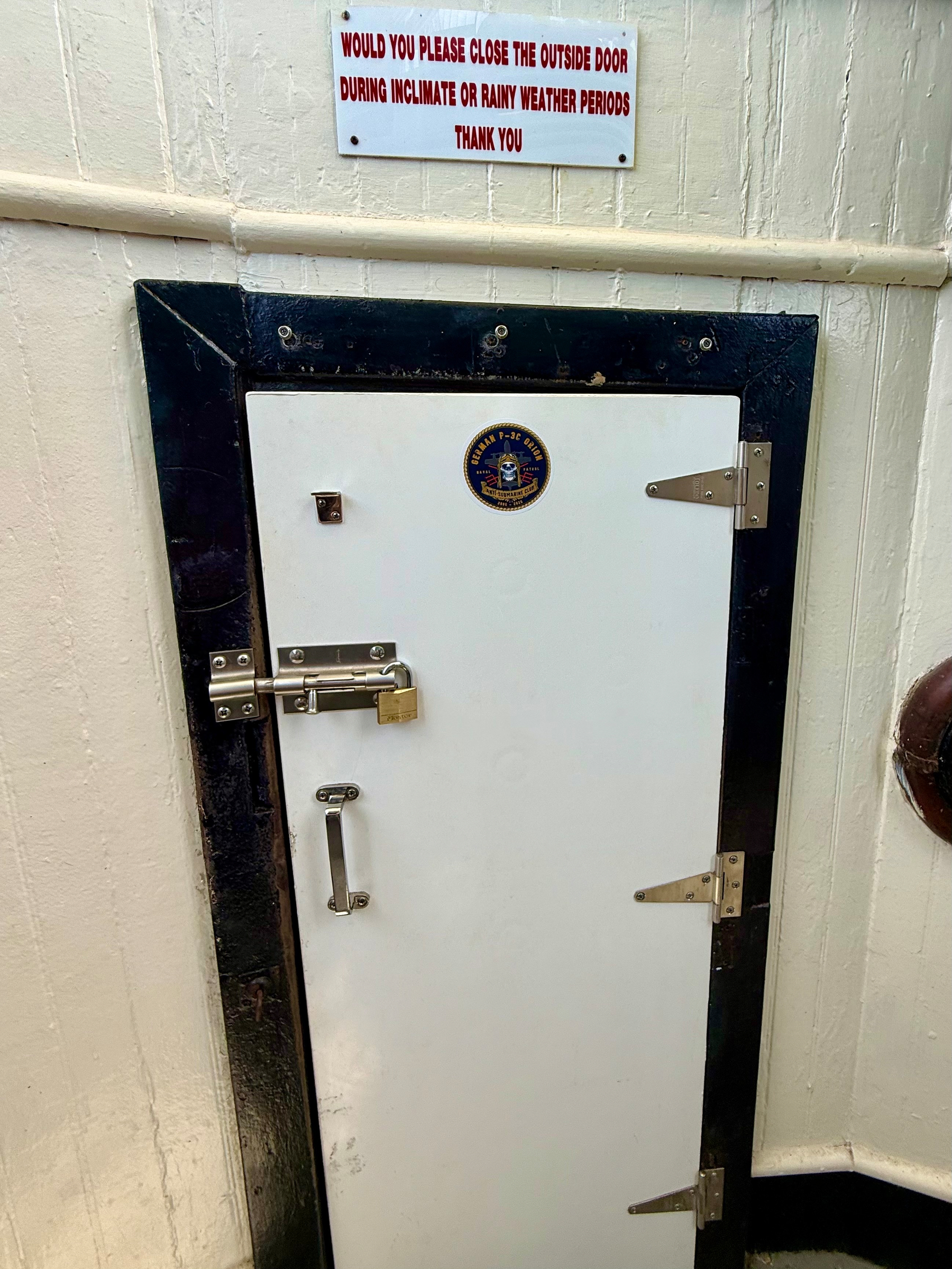 A small white metal door with a black frame and two silver hinges is secured by a silver latch with a brass padlock and has a silver handle below the latch. A circular emblem with a blue and gold design is displayed near the top center of the door. Above the door, a sign with red capital letters reads, “WOULD YOU PLEASE CLOSE THE OUTSIDE DOOR DURING INCLIMATE OR RAINY WEATHER PERIODS THANK YOU.” The surrounding wall is cream-coloured with a horizontal ridge running across it, and part of a circular brown object is visible on the right side.