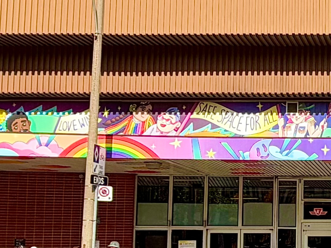 A colourful mural on the exterior of a building depicts a vibrant, inclusive scene. The mural features smiling cartoon-style characters of diverse appearances, some wearing rainbow-striped clothing and aviator uniforms. A rainbow arches across the bottom, surrounded by pink clouds, stars, and a cheerful, light-blue creature with a smiling face. The mural includes two banners: one on the left reading “LOVE WINS” and another on the right reading “SAFE SPACE FOR ALL.” The background is a mix of purple and blue tones with decorative stars and abstract shapes. Below the mural is a red brick wall with a windowed entrance, and a TTC (Toronto Transit Commission) sign is visible in the foreground attached to a pole.