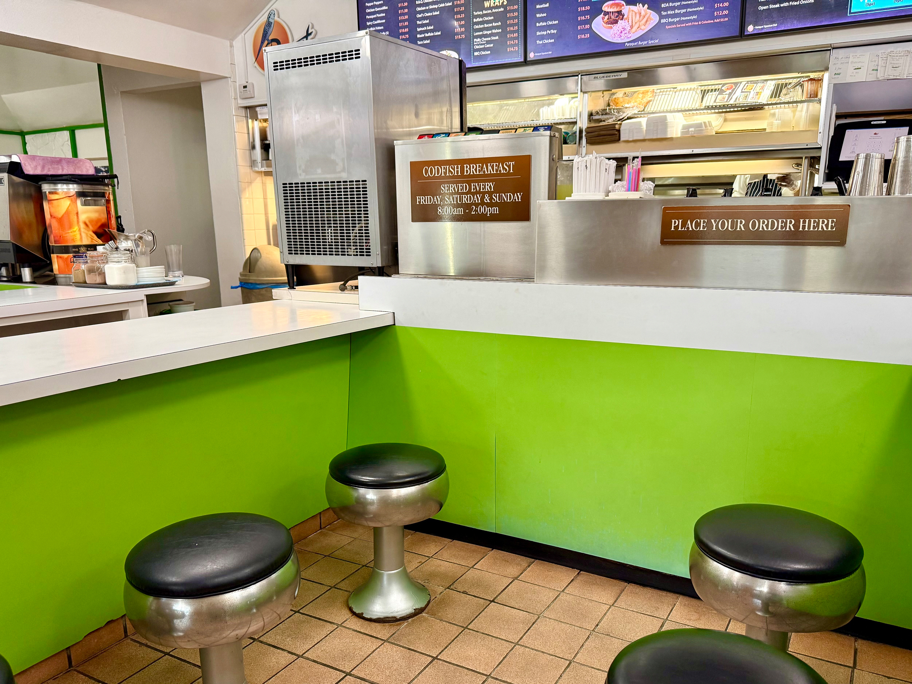 A small diner interior with bright green and white decor. Four round metal stools with black cushioned seats are arranged along a white counter with green paneling beneath. Behind the counter, there is a stainless steel service area with drink dispensers, straws, cups, and a food preparation station. A sign on the counter reads “PLACE YOUR ORDER HERE,” and another sign on a metal unit says “CODFISH BREAKFAST SERVED EVERY FRIDAY, SATURDAY & SUNDAY 8:00 a.m. – 2:00 p.m.” Above the counter, menus with images of food items are displayed. The floor is covered in light brown square tiles.
