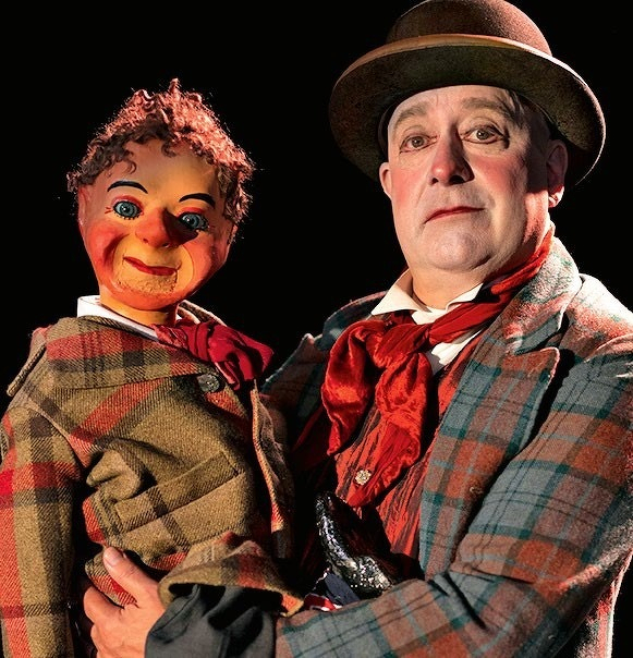 A ventriloquist is holding a puppet. The ventriloquist is wearing a plaid suit, a hat, and a red scarf, while the puppet is dressed similarly, also in plaid with a red tie. The puppet has a painted face with exaggerated features and curly hair. The background is dark, highlighting the characters.