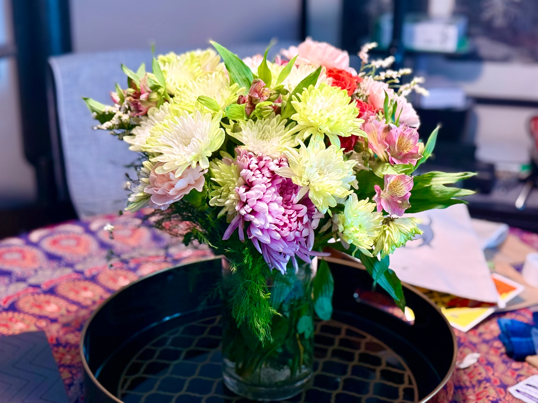 A glass vase filled with a vibrant bouquet of flowers, including pale yellow chrysanthemums, pink and lavender blooms, small red flowers, and green foliage. The bouquet is arranged in a lush, full display with some flowers slightly drooping over the edges. The vase sits on a black tray with a gold geometric pattern, placed on a table covered with a colourful, patterned cloth featuring red, blue, and purple tones. In the background, there are blurred household items, including papers and a white napkin, creating a casual indoor setting.