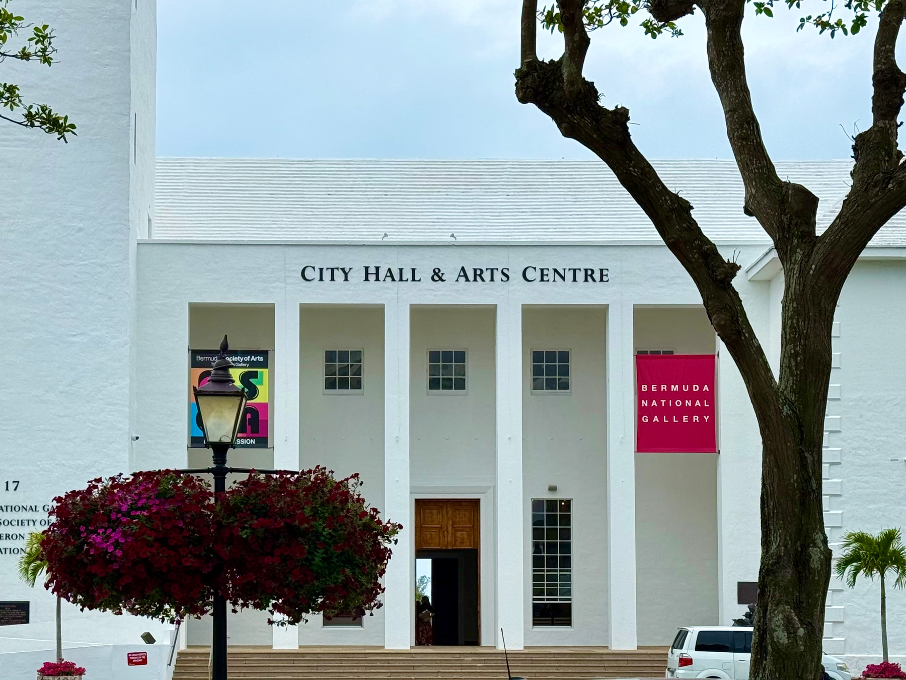 A white building with a gabled roof and multiple tall rectangular windows, identified as City Hall & Arts Centre. The entrance has a wooden double door at the top of a wide set of steps, flanked by four large columns. On the left side, a colourful poster for the Bermuda Society of Arts is displayed, and on the right side, a bright red banner reads "Bermuda National Gallery." A black streetlamp with two large hanging baskets of red and purple flowers stands in front of the building. A tree with a thick trunk and spreading branches partially frames the right side of the image, and a white vehicle is parked near the entrance. The sky is overcast, and there are small palm trees in the foreground.