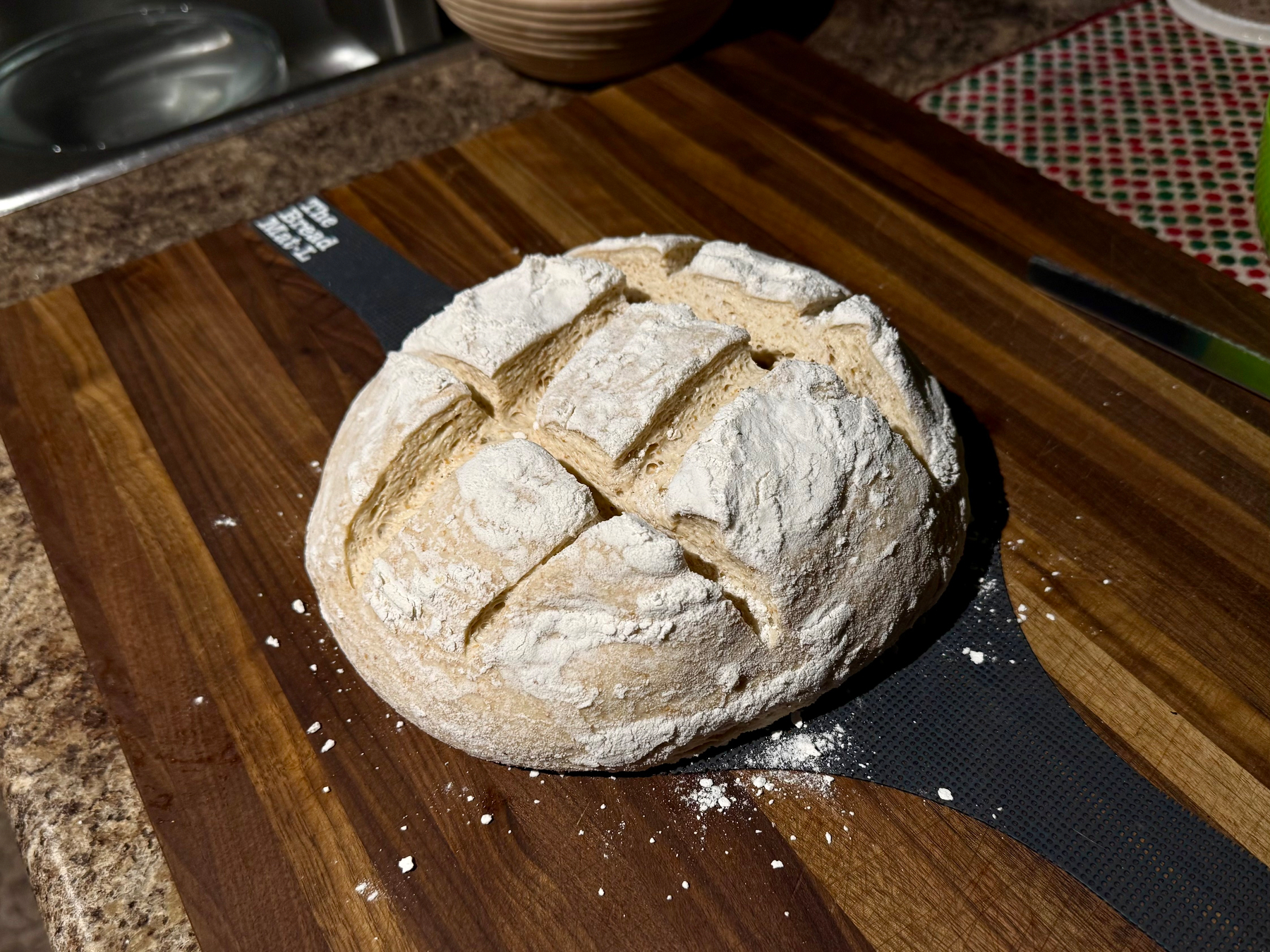 A round loaf of rustic bread sits on a wooden cutting board with a dark inlay strip. The bread is dusted with flour and has a golden-brown crust with deep square-shaped scoring on top, creating a quilted pattern. Some flour is scattered on the board around the loaf. In the background, part of a kitchen counter with a sink, a patterned mat, a serrated knife, and a wooden bowl is visible.