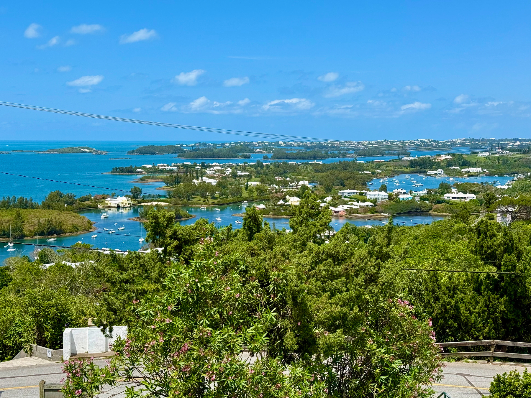 A scenic coastal landscape with a bright blue sky dotted with small clouds. The view overlooks turquoise waters with numerous small islands and inlets, surrounded by lush green vegetation. White-roofed houses are scattered along the shoreline, and several boats are anchored in the calm bays. In the foreground, flowering green shrubs and trees line a narrow road with a stone wall and wooden guardrail. Power lines stretch horizontally across the upper part of the scene.