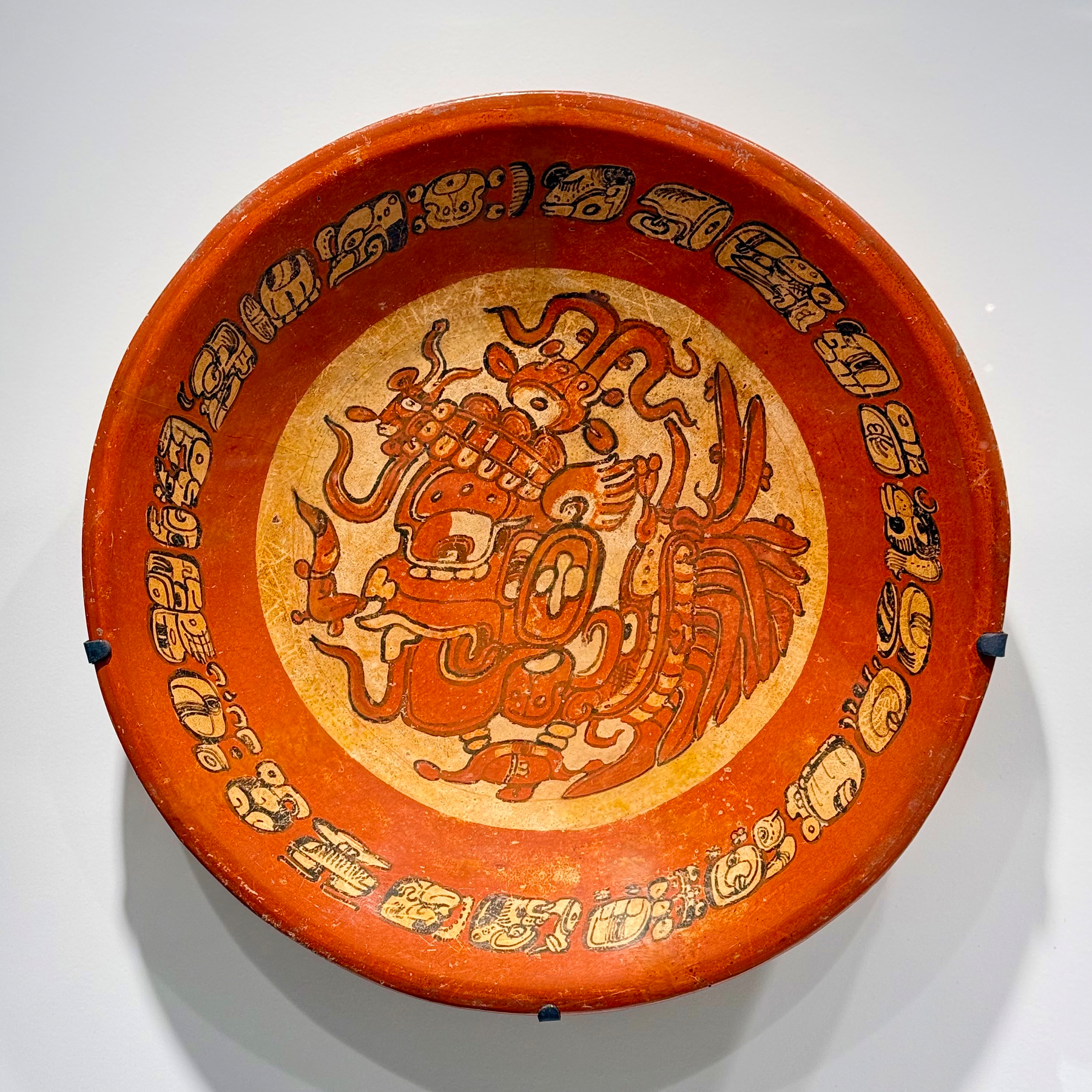 A round, intricately decorated ceramic plate with a reddish-brown border and a central design depicting a stylized mythical figure or creature. The border features intricate, repeating glyph-like patterns. The main artwork inside the plate exhibits detailed line work and appears to be influenced by ancient or indigenous art styles. The plate is mounted and displayed against a light background.