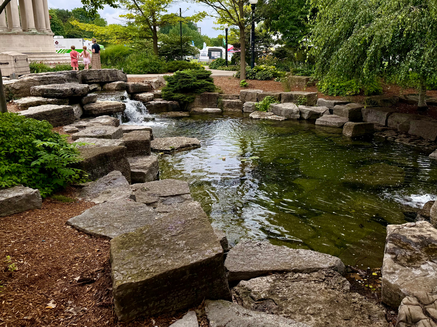A small, rocky pond with a waterfall feature is surrounded by greenery and a few people standing near a stone structure in a park setting.