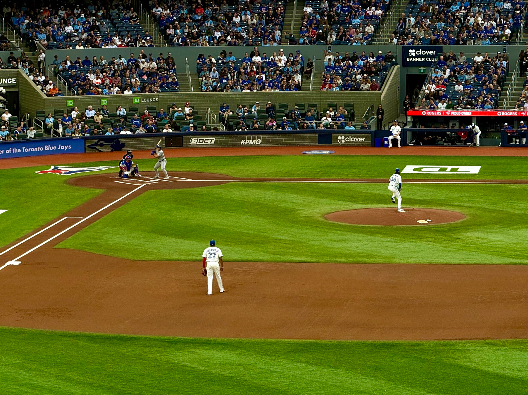 A professional baseball game in progress at a stadium with a large crowd in the stands. The pitcher in a white uniform is mid-delivery on the pitcher’s mound, while a batter in a grey uniform prepares to swing at home plate. Behind the batter, the catcher is crouched in position, and an umpire is standing behind the catcher. A player wearing a white uniform with the number 27 and the name "Guerrero Jr." stands on the infield near third base. The field is well-manicured with bright green grass and reddish-brown dirt. Fans fill the lower and upper seating areas, with some sections appearing more densely packed than others. Advertisements for brands like KPMG, SBET, clover, and Rogers are visible around the field, alongside a banner that reads “Home of the Toronto Blue Jays.”