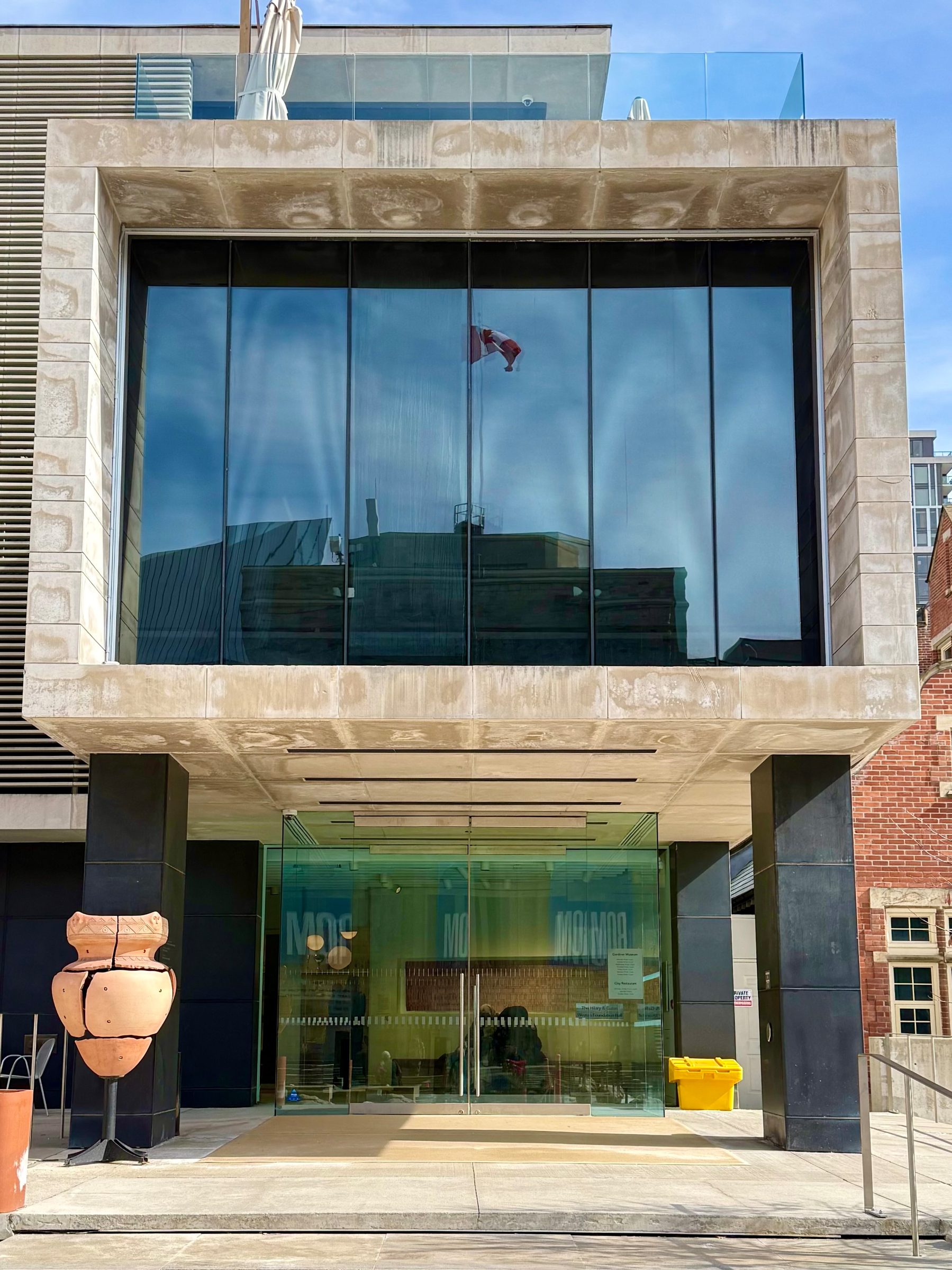 A modern building entrance with large glass windows reflecting a Canadian flag in the background. The facade is framed with a beige stone structure. A large ceramic sculpture stands to the left of the entrance, while a yellow bin is visible on the right. The sky is clear and sunny.