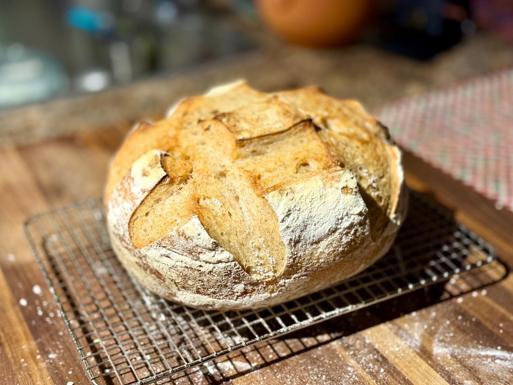 A freshly baked round loaf of artisan bread rests on a cooling rack atop a wooden countertop. The bread has a golden-brown crust with a rustic, flour-dusted surface and deep scoring in a cross pattern, allowing the crust to open and create crisp ridges. In the background, the kitchen counter is slightly blurred, with a patterned cloth to the right and various kitchen items out of focus, emphasizing the warm, homemade feel of the scene.