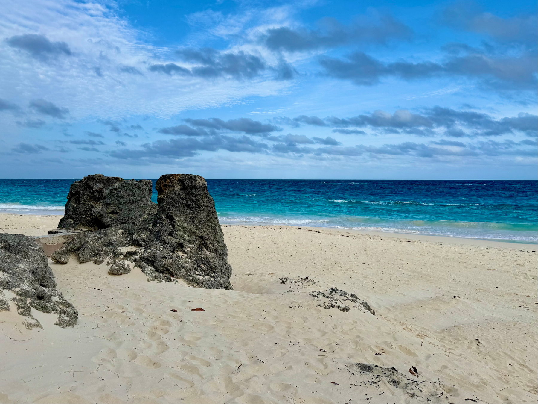 A sandy beach with light beige sand stretches toward the turquoise and deep blue ocean, meeting the horizon under a partly cloudy sky. A large weathered rock formation sits in the foreground, split into two uneven sections, with smaller rocks and scattered debris embedded in the sand around it. The gentle waves lap against the shore, and the clouds vary from fluffy white to darker grey, creating a serene and natural coastal scene.