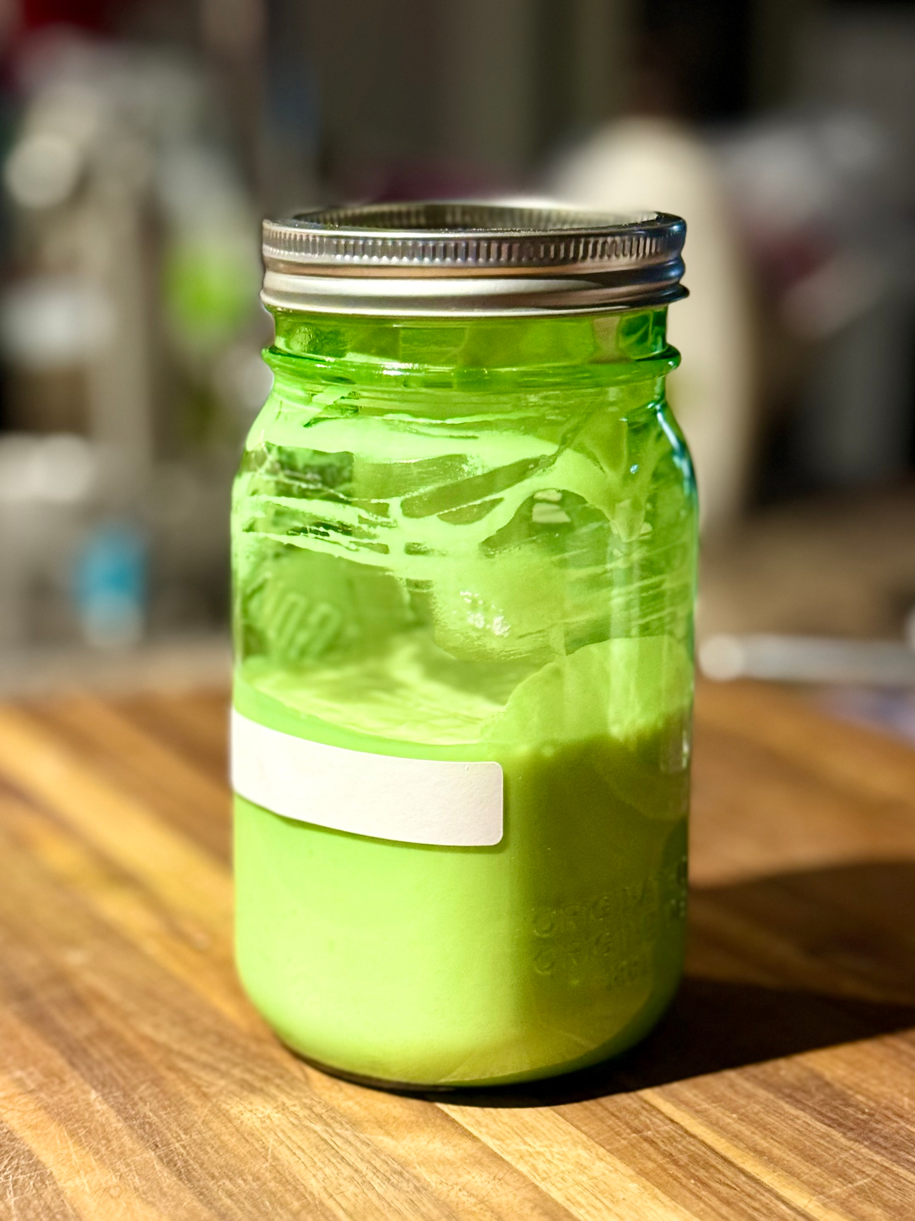 A glass jar with a metal lid filled with sourdough starter sits on a wooden surface. The jar has a blank white label on the front. The background is softly blurred, bringing focus to the jar's vibrant contents.