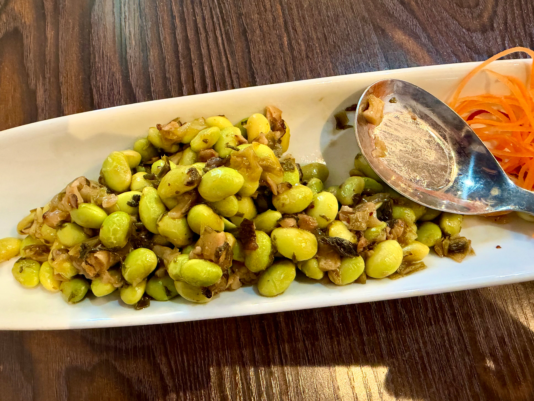 A dish of cooked green soybeans mixed with small pieces of brown mushrooms is served on a rectangular white plate with a silver spoon and a garnish of shredded carrots on the side. A white rectangular plate on a wooden table holds a serving of cooked edamame beans mixed with small pieces of chopped vegetables, possibly onions and greens. A large metal spoon rests on the right side of the plate, next to a small garnish of thinly sliced, curled orange carrots. The edamame mixture appears lightly seasoned with visible oil and browned bits from cooking.