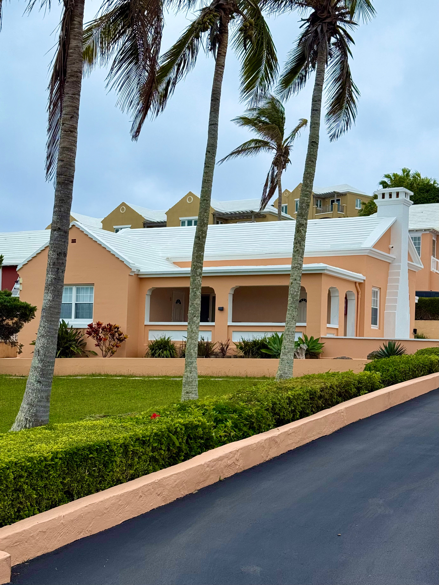 A peach-coloured house with white trim and a white stepped roof sits behind a neatly maintained lawn, bordered by a low hedge and a peach retaining wall. Tall palm trees sway in the wind in the foreground under a cloudy, overcast sky. The house features a covered front porch with arched openings and a white chimney on the right side. In the background, tan-coloured buildings with matching white roofs are visible. The driveway in the foreground is paved in dark asphalt, contrasting with the greenery and pastel tones of the property.