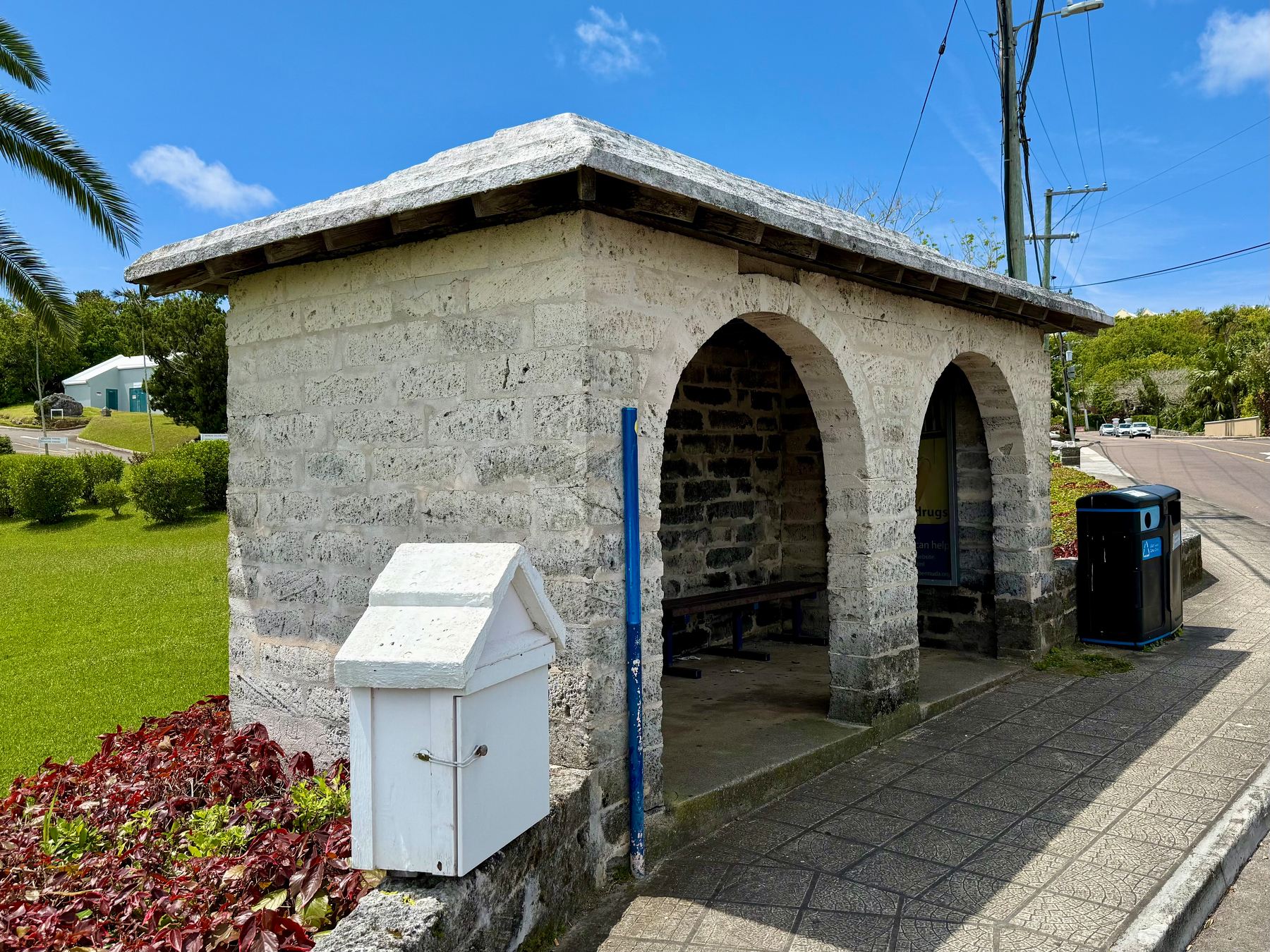 A small stone bus shelter with two arched openings sits beside a paved sidewalk on a sunny day. The shelter is built from light grey stone blocks with a sloped white roof, and inside there is a simple wooden bench. To the left, a small white box with a pitched roof is mounted on the stone wall near a bed of red and green plants. On the right side of the shelter, a black and blue public garbage and recycling bin stands by the sidewalk. Behind the shelter, a manicured lawn stretches toward a light blue building with a driveway and trimmed bushes. A clear blue sky with a few wispy clouds and palm tree fronds completes the scene.