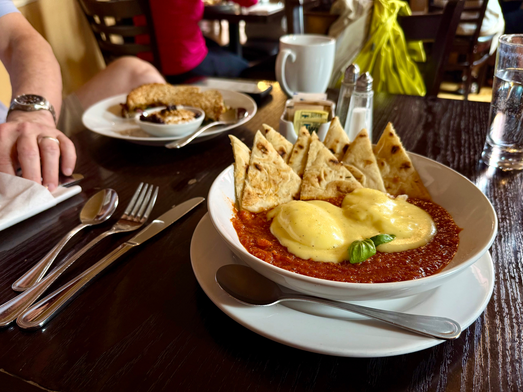A plate of eggs Benedict topped with hollandaise sauce is served with pita bread triangles on a table set with utensils and a glass of water in a restaurant setting.