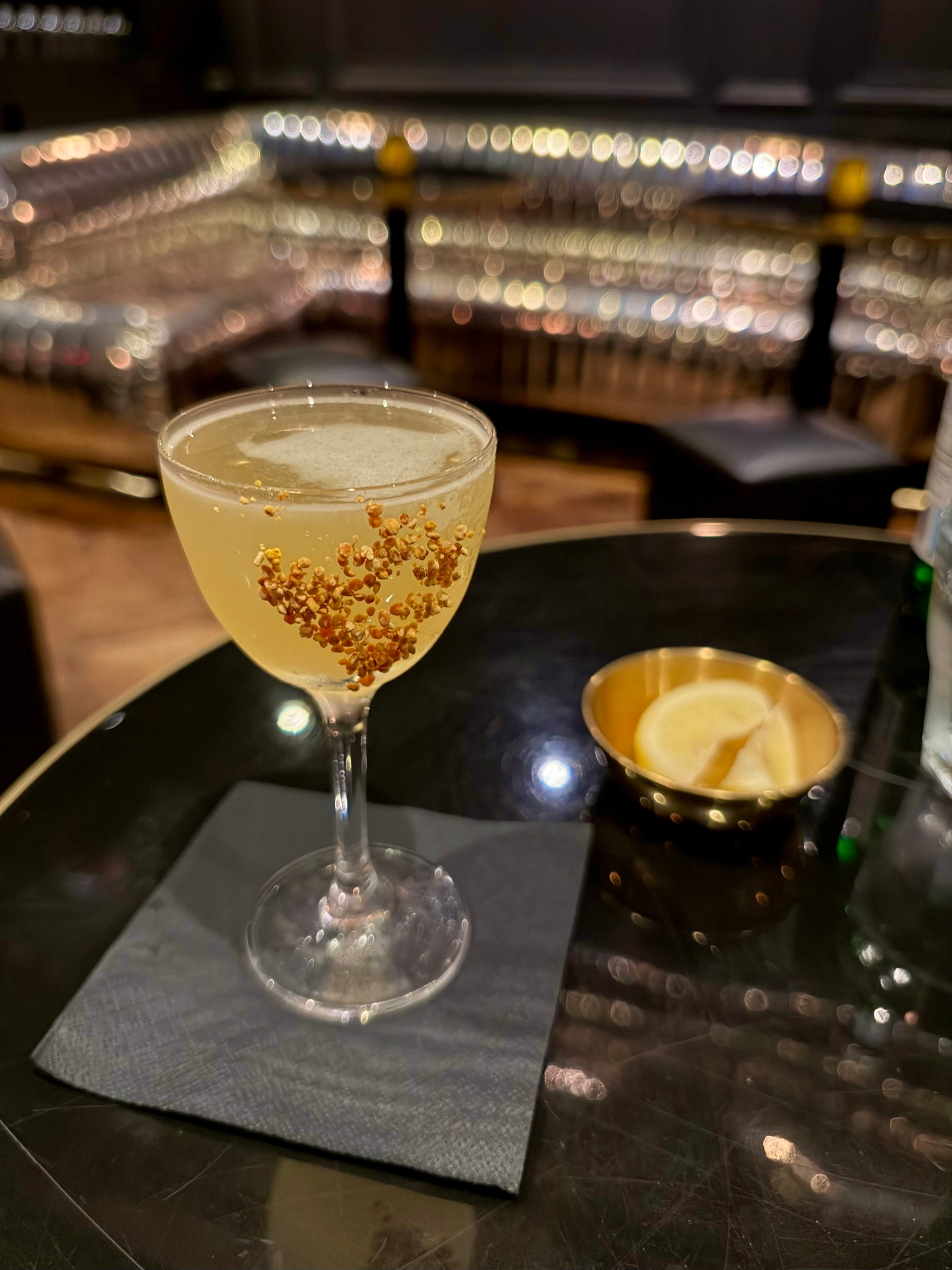 A clear cocktail glass sits on a dark reflective table with a black napkin underneath. The glass contains a pale yellow drink garnished with small golden spherical seeds floating inside. Behind the drink, a small round metal bowl holds lemon wedges. The background shows a shiny, metallic, curved bench with a high-gloss surface, reflecting warm ambient lighting, and a few blurred stools and tables. The overall atmosphere is dim and elegant, with a focus on the drink in the foreground.
