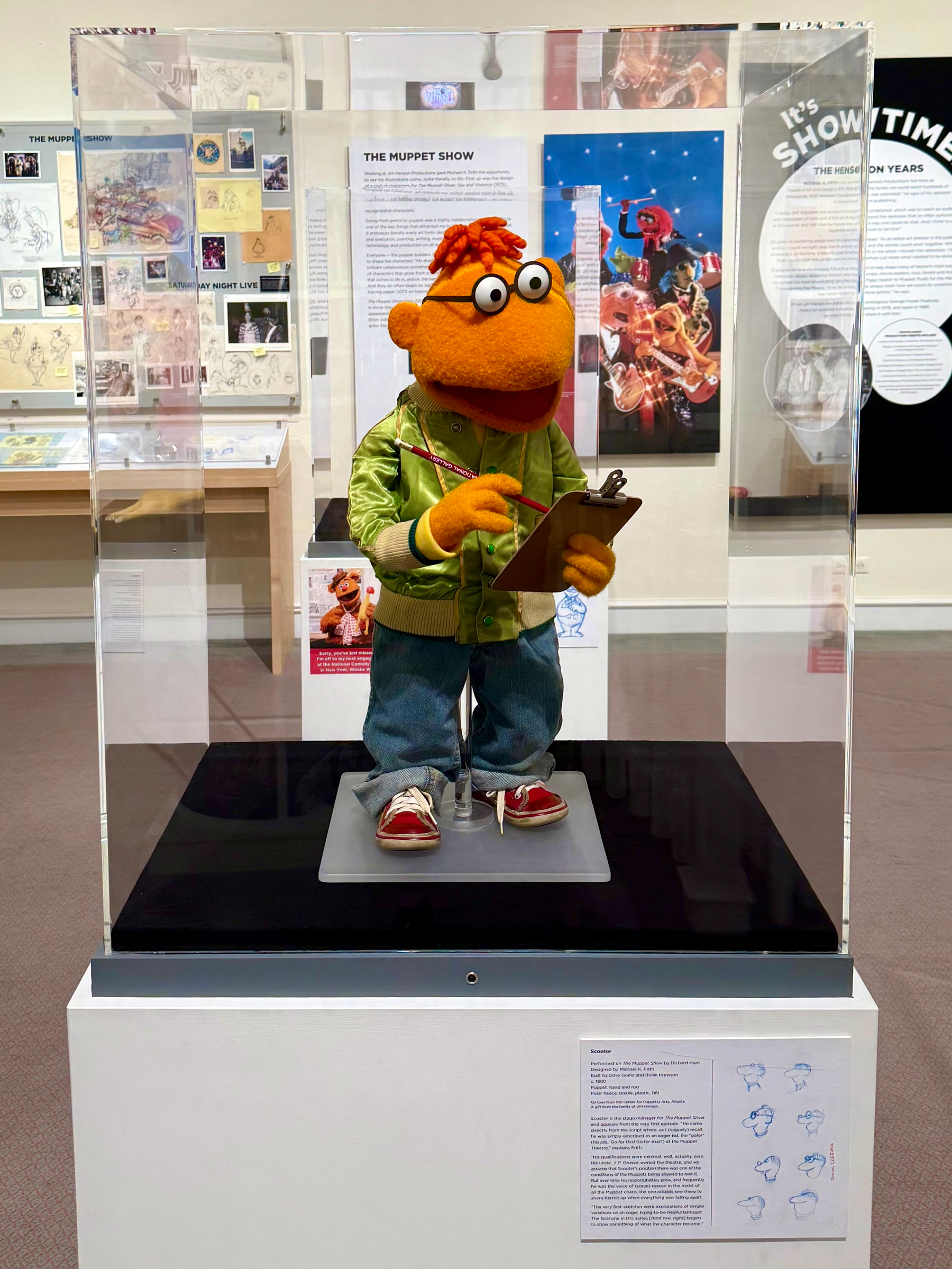 A museum display features a Muppet character named Scooter standing inside a clear protective case. Scooter is an orange puppet with orange hair, large round white eyes with black pupils, and a wide smiling mouth. He is wearing a shiny green satin jacket with yellow cuffs, blue jeans, and red sneakers with white soles. He is holding a clipboard and a red pencil, appearing as if taking notes. The case is mounted on a black platform with a white pedestal underneath. The background shows a museum exhibit with posters, sketches, and photos related to The Muppet Show. Behind the display, a large panel with the title “THE MUPPET SHOW” provides information, and another panel shows colourful images of Muppet characters performing. To the right of the pedestal, a descriptive plaque includes a paragraph of text about Scooter along with several blue sketches of his head drawn in different angles and expressions.