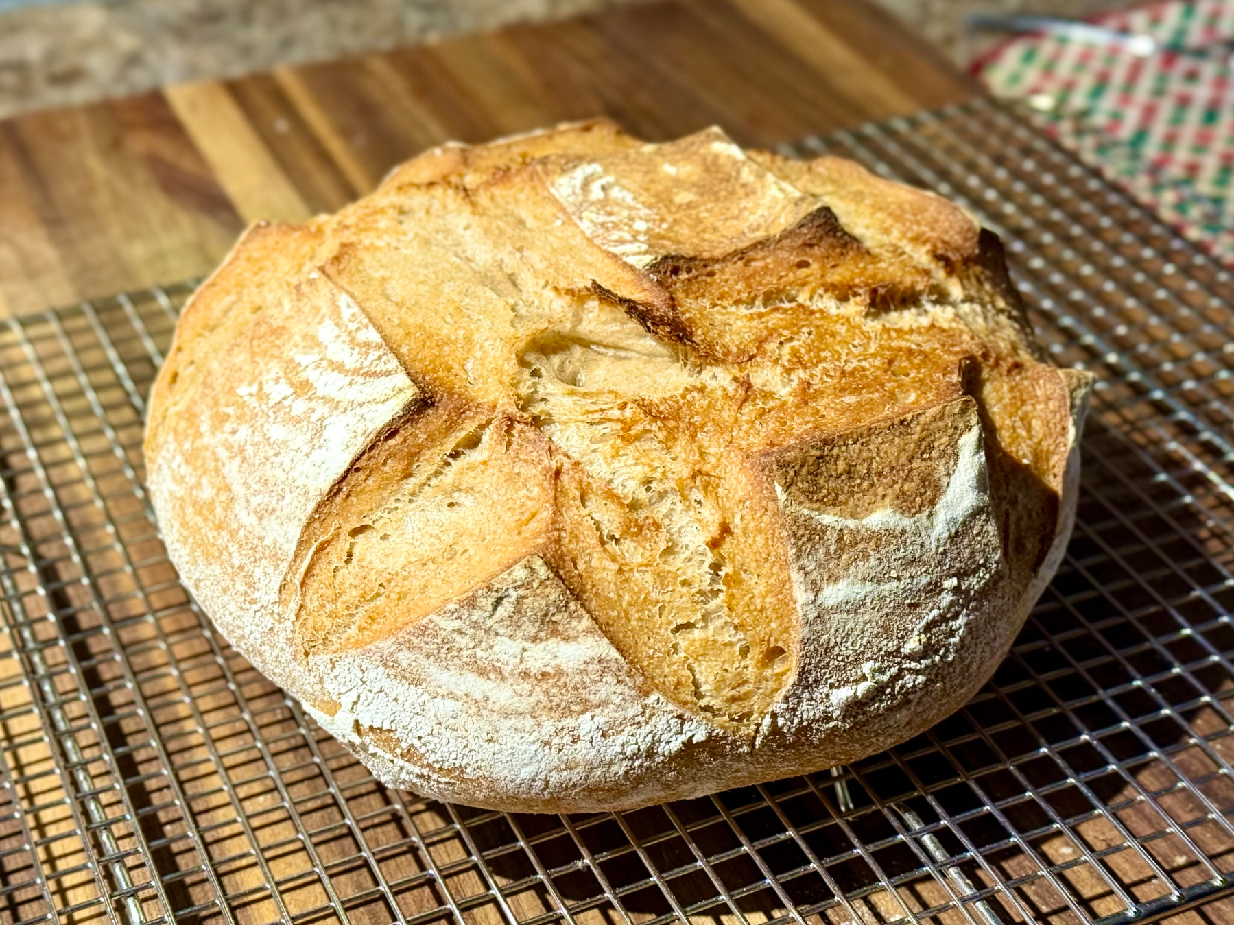 A freshly baked round loaf of bread with a golden-brown crust and detailed scoring patterns, resting on a cooling rack. The bread has a rustic appearance and is set against a wooden surface.