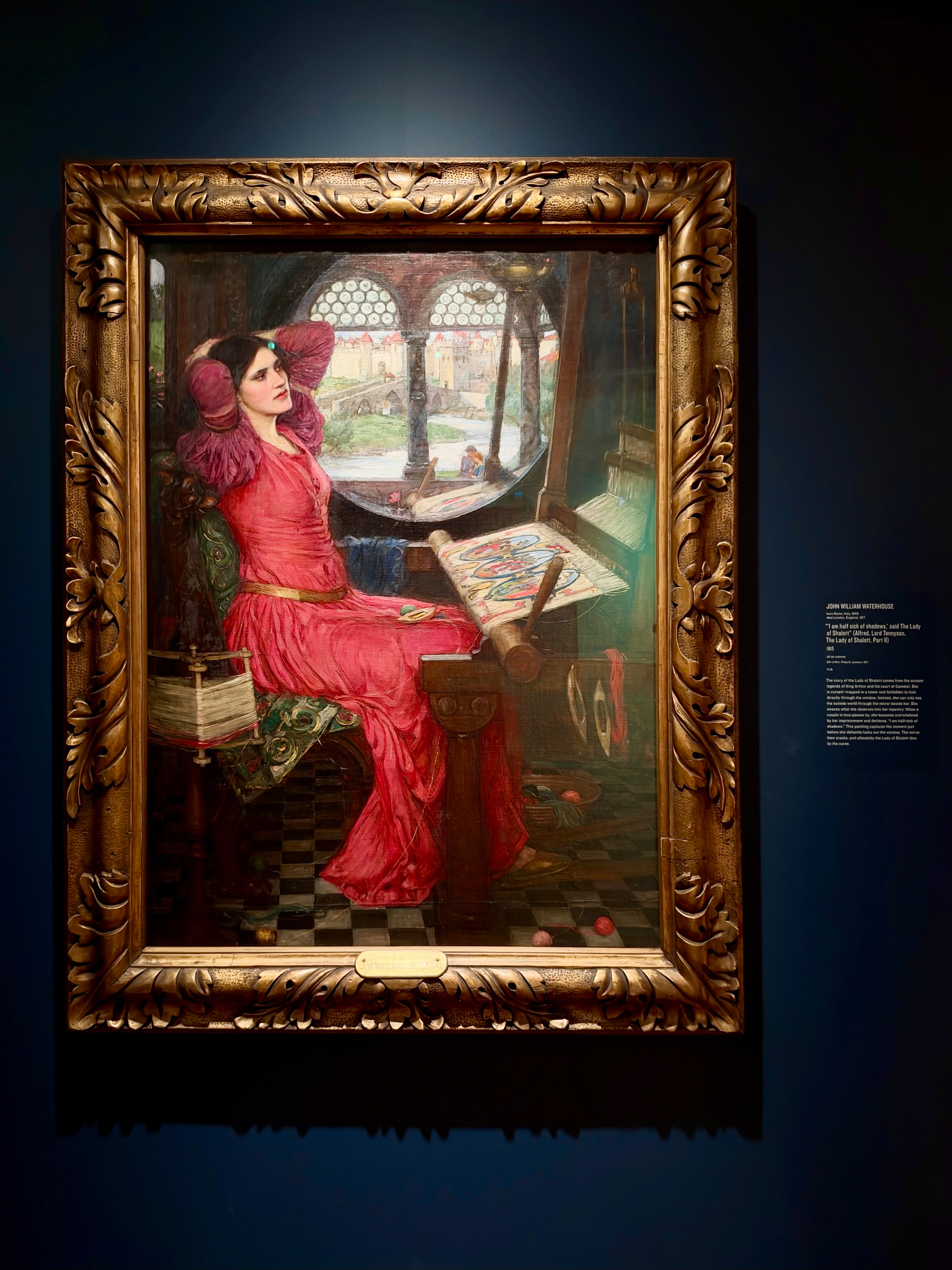 A framed painting displayed against a dark blue wall in a museum or gallery setting. The ornate gold frame features intricate leaf and scroll designs. The painting depicts a woman dressed in a long red gown with puffed sleeves, sitting on a green cushioned chair adorned with embroidered patterns. She leans back with her arms raised, gazing to the side with a pensive expression, framed by dark hair and a fair complexion. In front of her is a large wooden loom holding a partially completed tapestry with colourful threads. The floor is made of black and dark grey checkered tiles, and several balls of yarn are scattered nearby. Behind her, a circular window with multiple small glass panes reveals a bright, sunlit exterior with a river, stone bridge, trees, and a distant castle-like structure with red rooftops. On the right side of the wall, there is a small label with text identifying the painting and providing information about the artist and work.